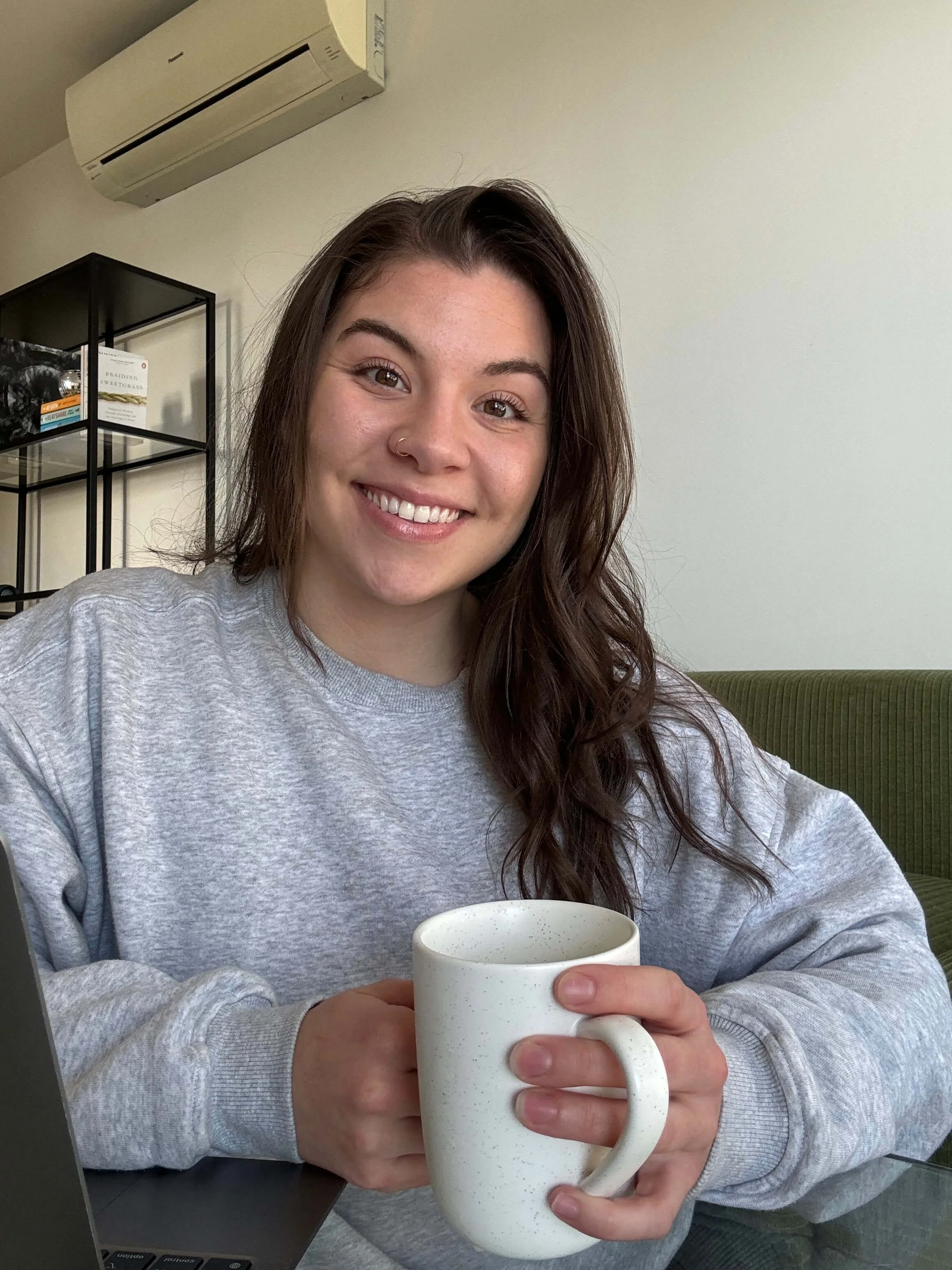 Emma Lapierre, Accredited Mental Health Social Worker, therapist for teens and young adults. A woman with long brown hair, smiling, holding a white speckled mug, sitting at a desk with a laptop, in a room with white walls, and a green couch.