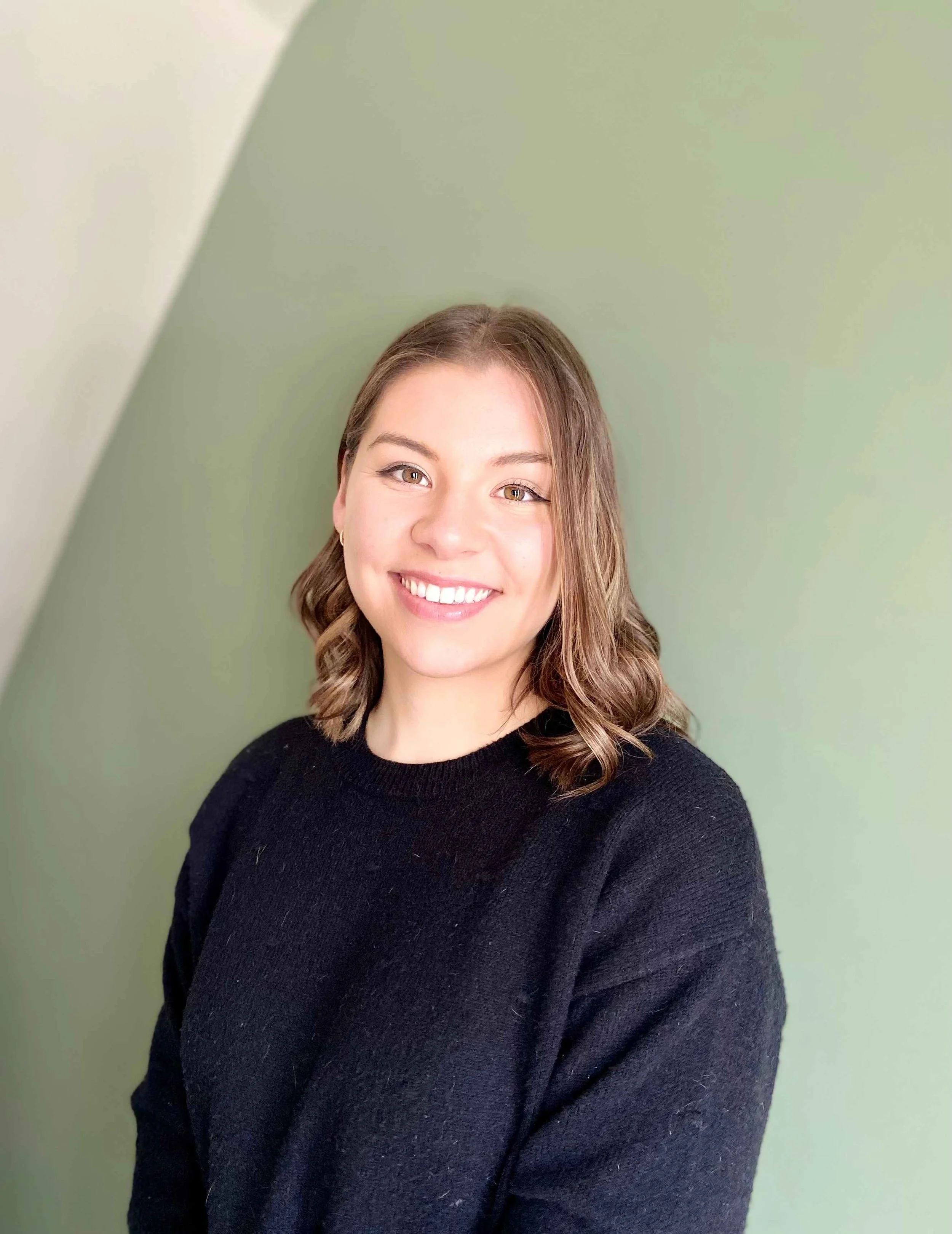 A young woman with shoulder-length wavy brown hair smiling, wearing a black sweater, standing against a two-tone green wall.