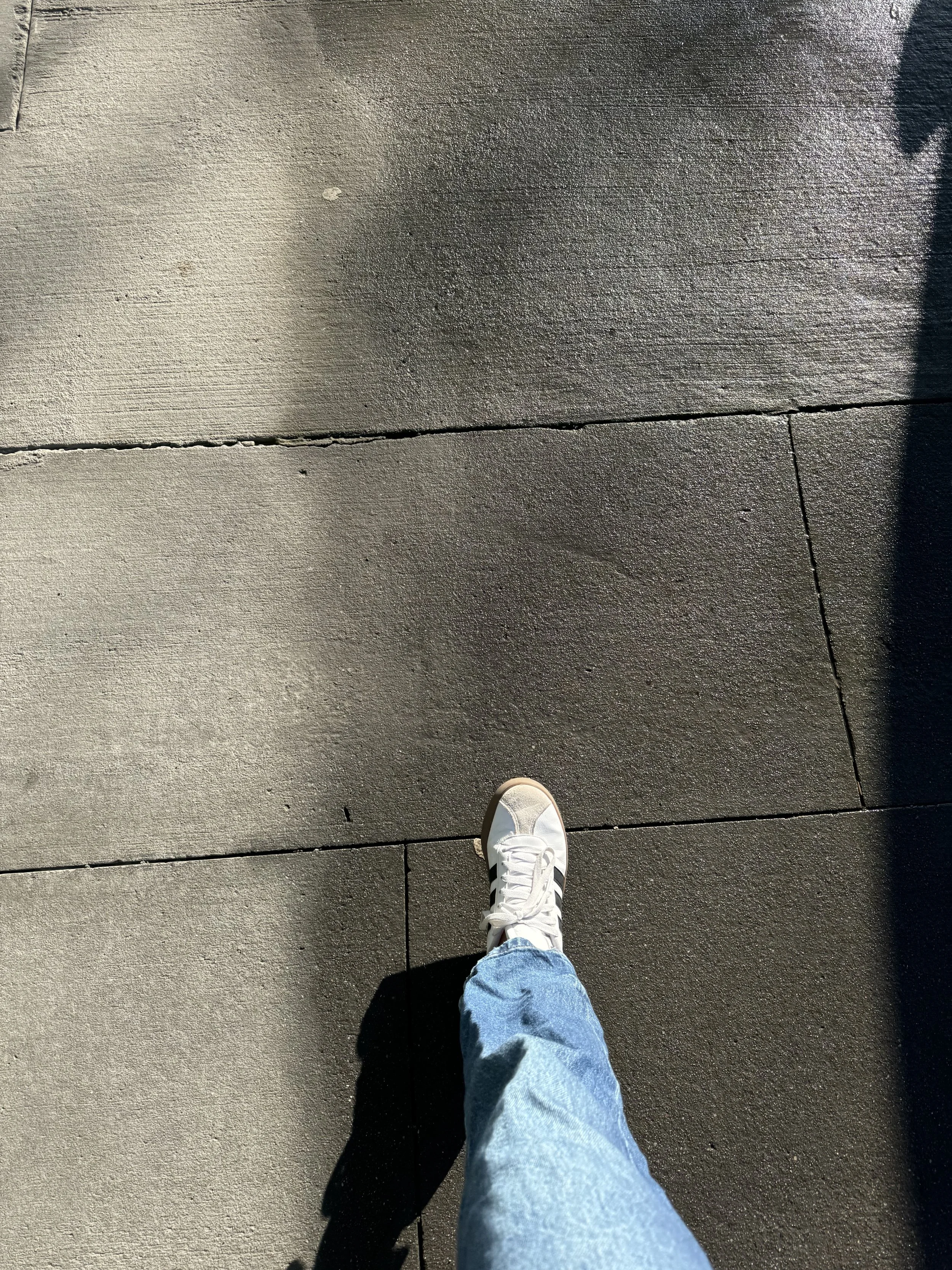 A person's foot in a white sneaker and light blue jeans walking on concrete sidewalk with sunlight and shadows.