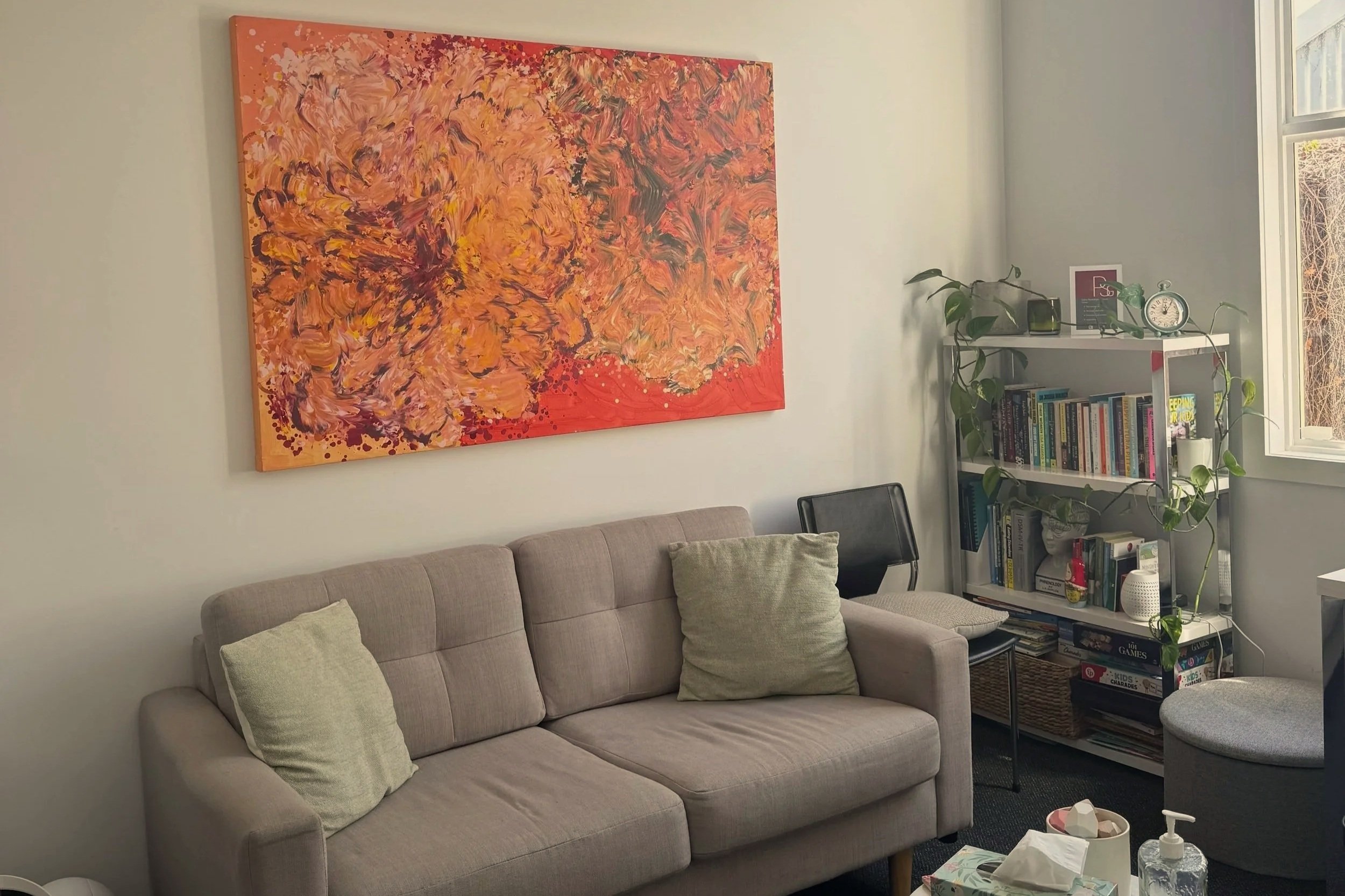 Therapy office with beige sofa, green pillows, abstract orange and pink floral painting on the wall, black chair, bookshelf with books, plants, clock, and window.