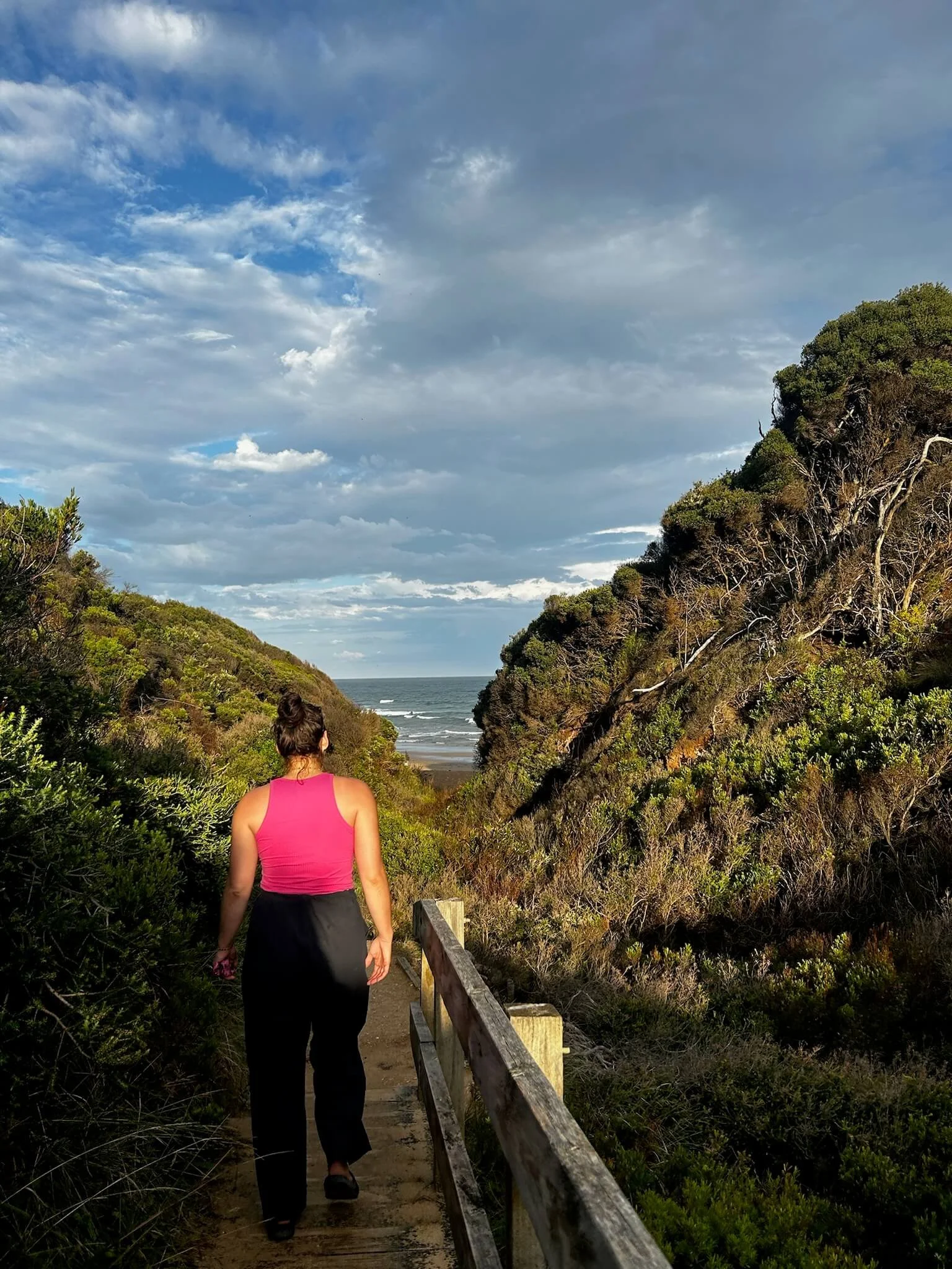 Therapy for teens, young adults, and athletes. A woman with a pink tank top and black pants walking along a wooden pathway through a natural landscape towards the ocean, with a partly cloudy sky above.