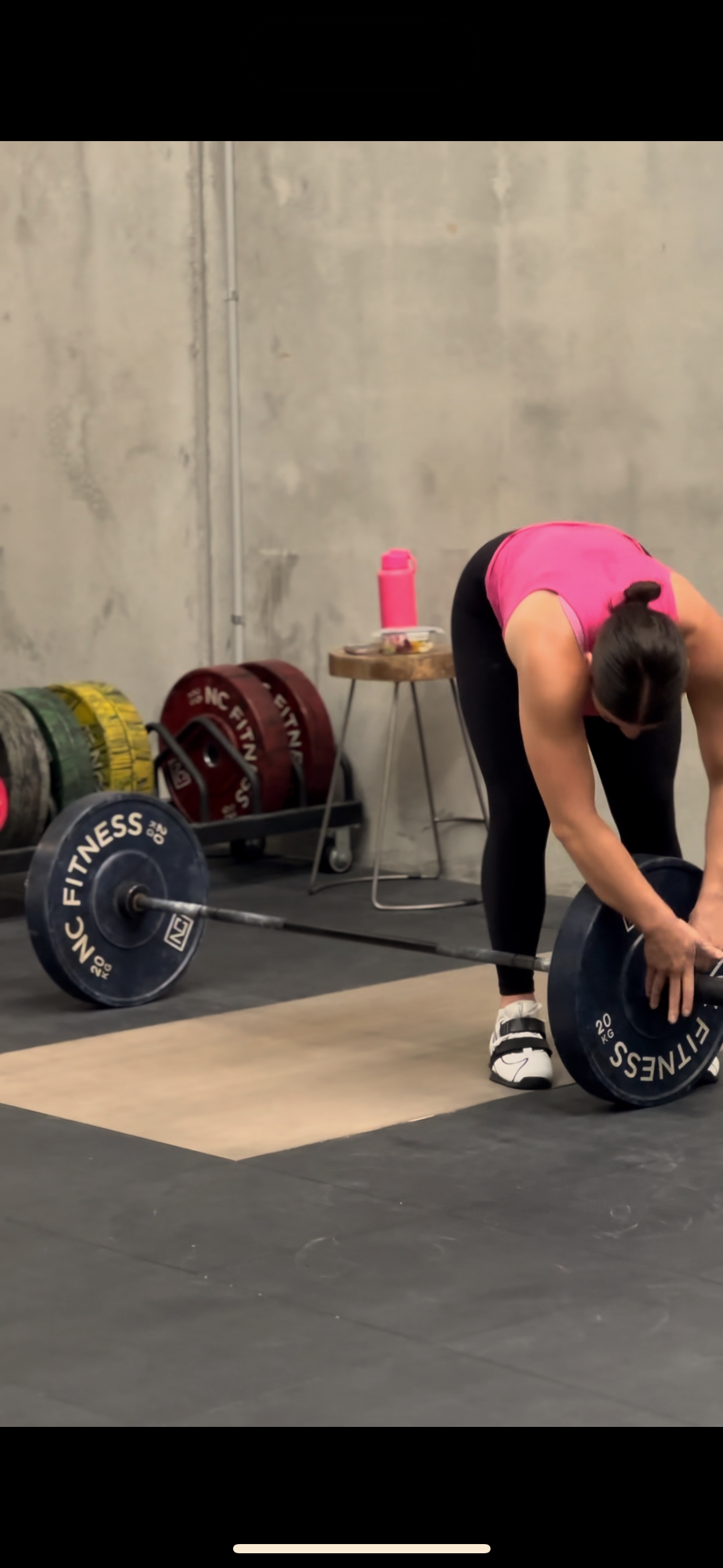 A woman in a pink tank top and black leggings preparing to lift a barbell with weights at a gym.