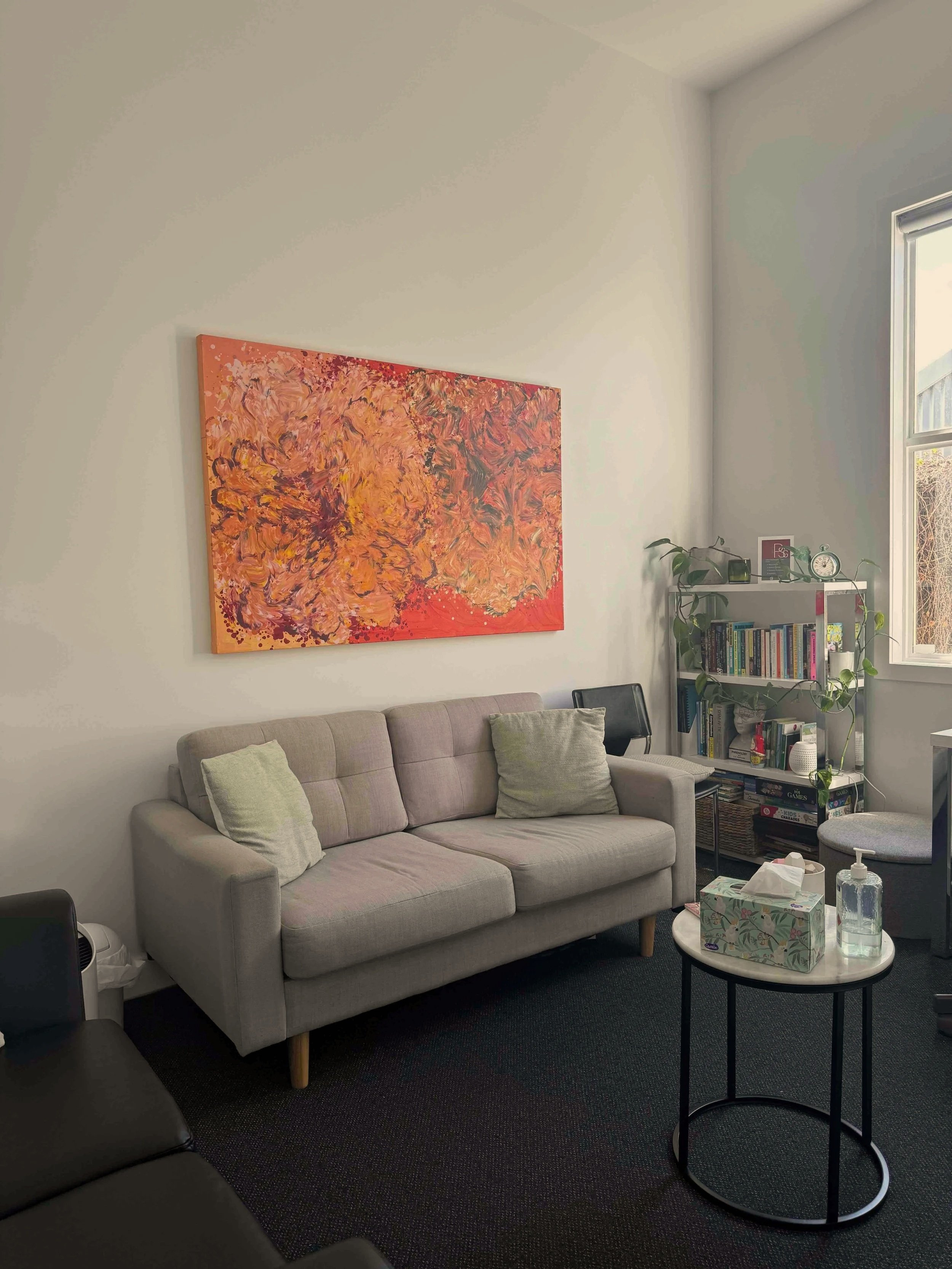 Therapy for teens and young adults. Living room with a beige couch, a colourful abstract painting on the wall, a bookshelf with plants, tissues, and hand sanitizer on a round table.
