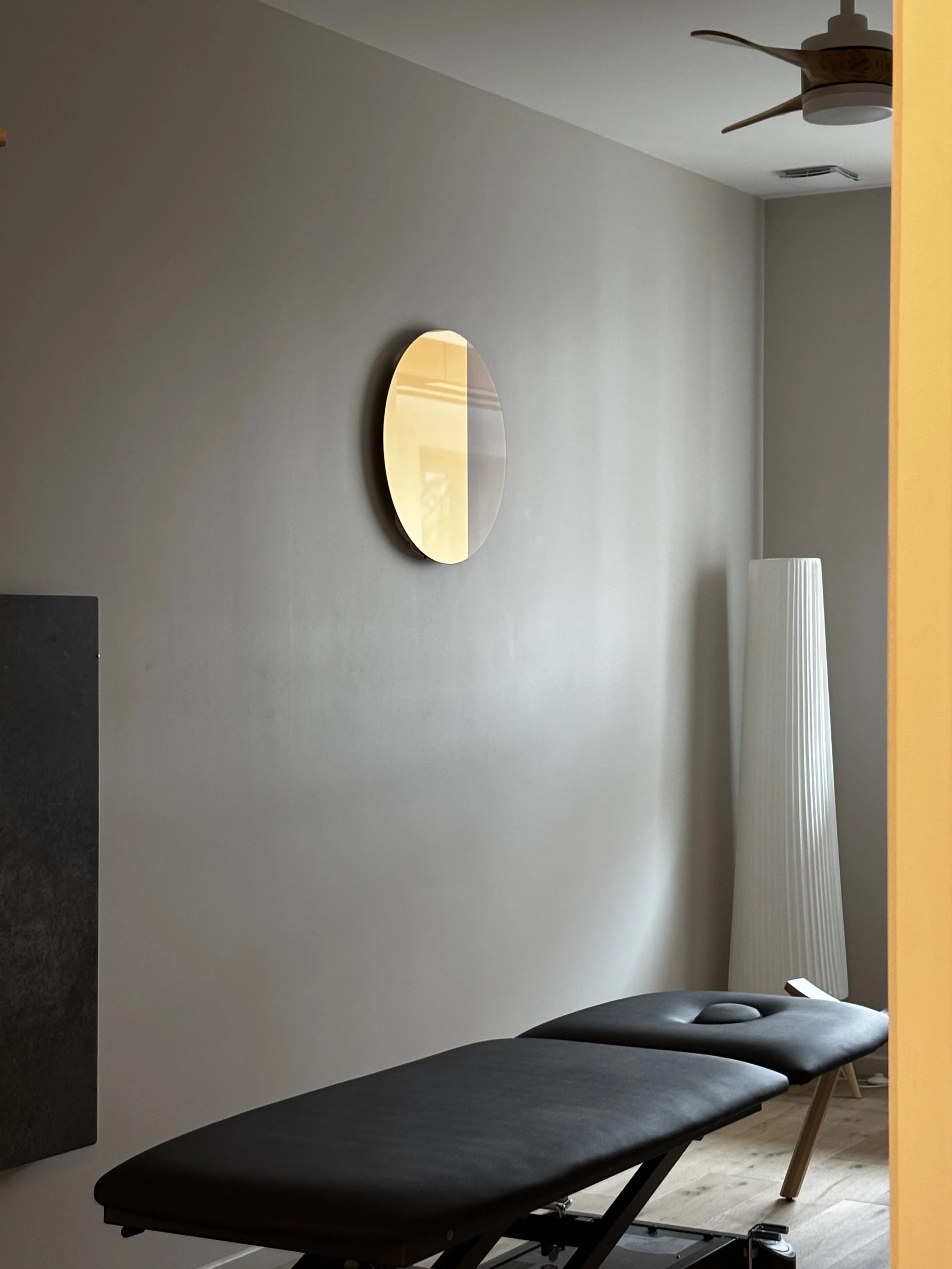 Une salle de thérapie ou de soins avec une table de massage ou d'examen noir, un miroir rond au mur, une lampe à pied blanche et un ventilateur au plafond.