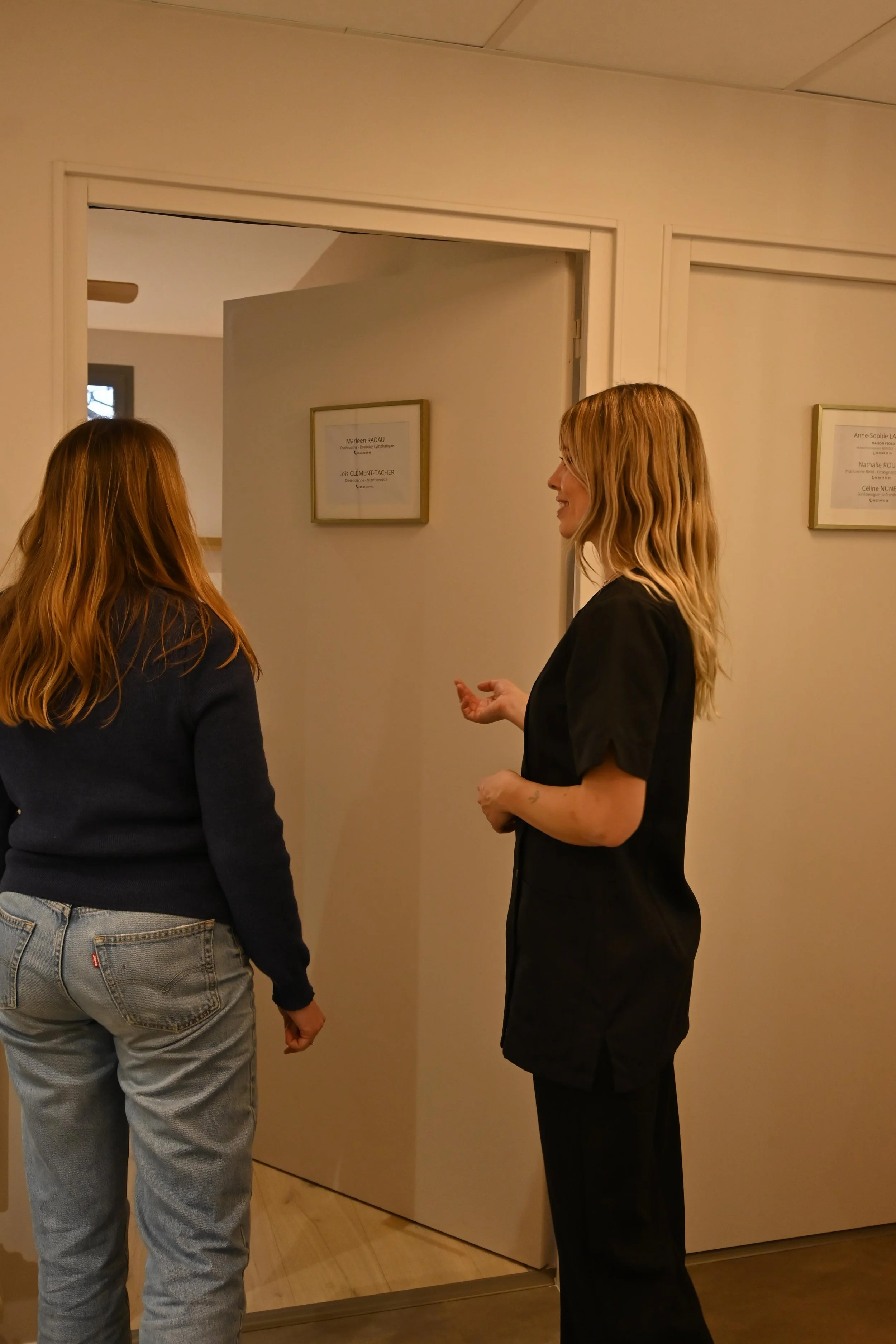 Deux femmes discutent dans un couloir d'un professionnel de santé, avec des plaques nominatives accrochées aux portes.