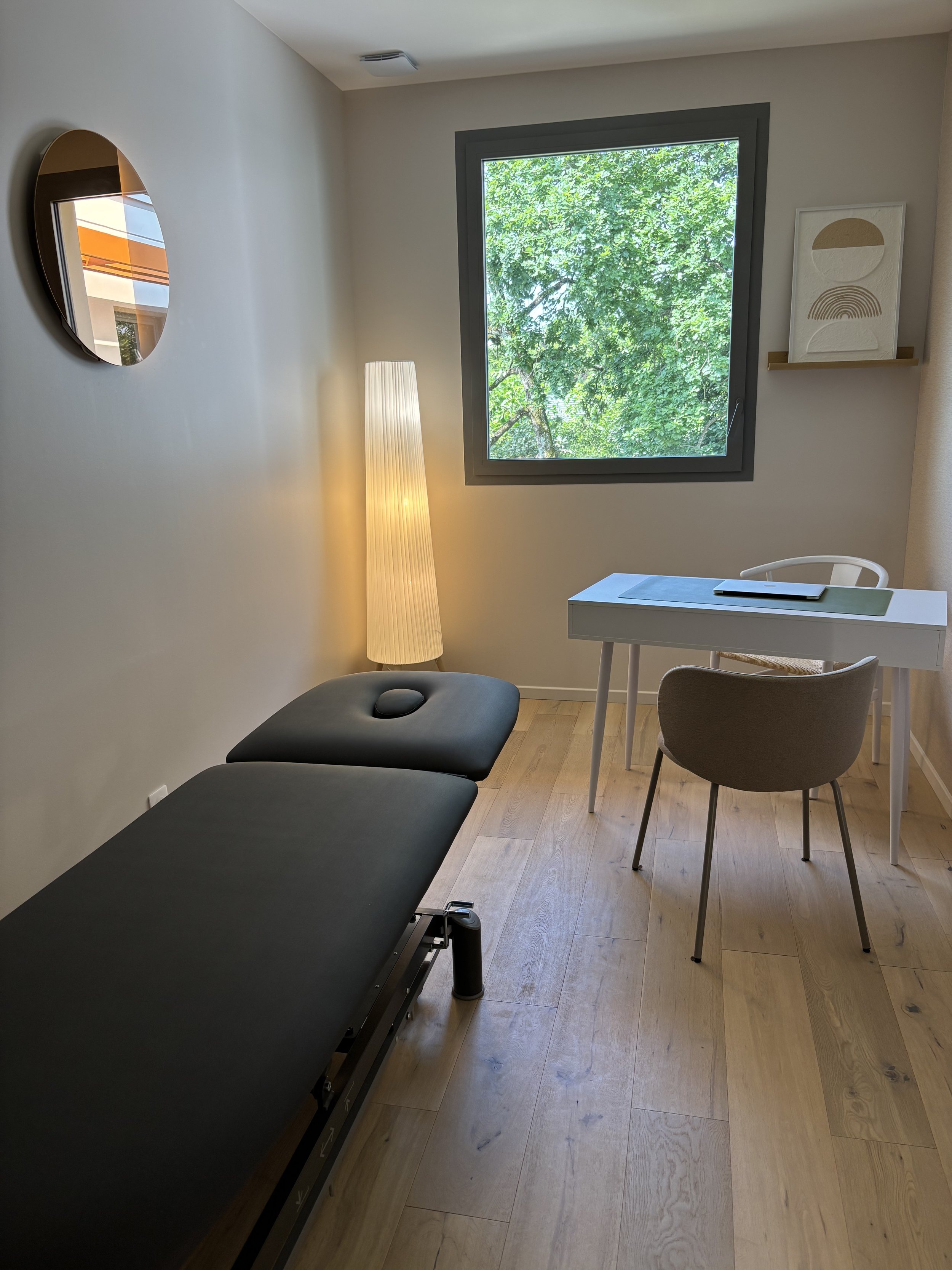 Une salle de consultation avec une table de massage noire, une lampe à lampadaire, un bureau blanc avec une chaise, un mur avec un miroir rond, une fenêtre avec une vue sur des arbres, et des décorations minimalistes.