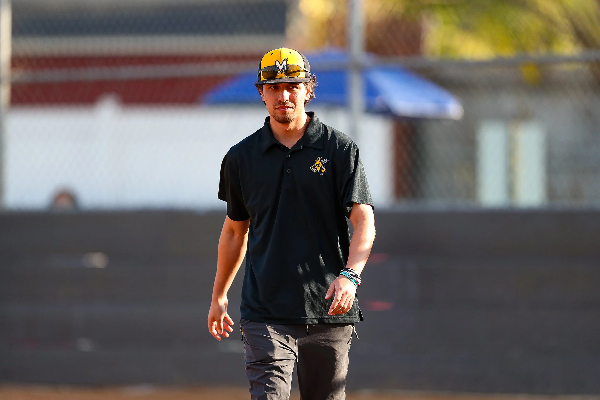 Coach Aaron wearing sunglasses, a black polo shirt with a sports team logo, gray pants, and a baseball cap with a yellow and black design walking on a baseball field.