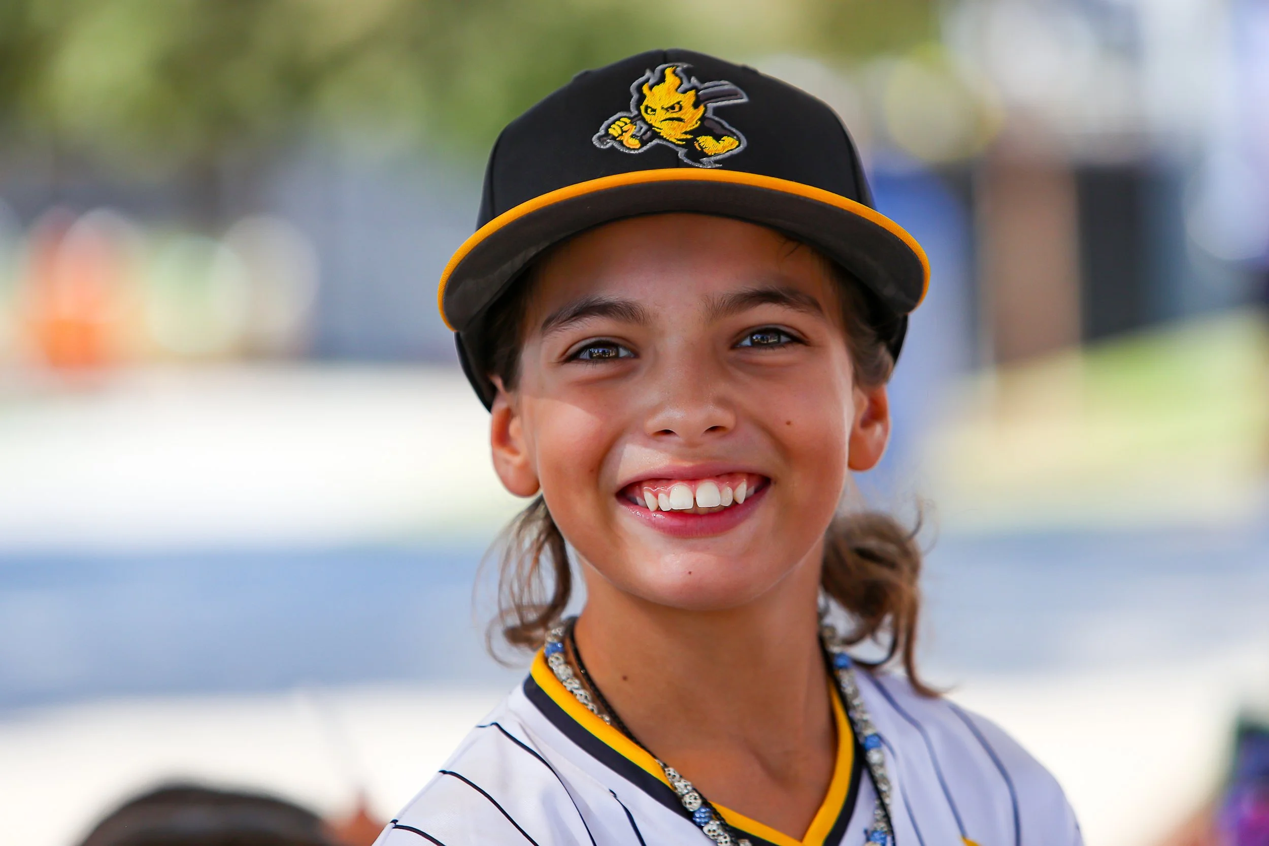 A young boy smiling, wearing a baseball cap with a lightning mascot and a white jersey with black stripes, outdoors on a sunny day.