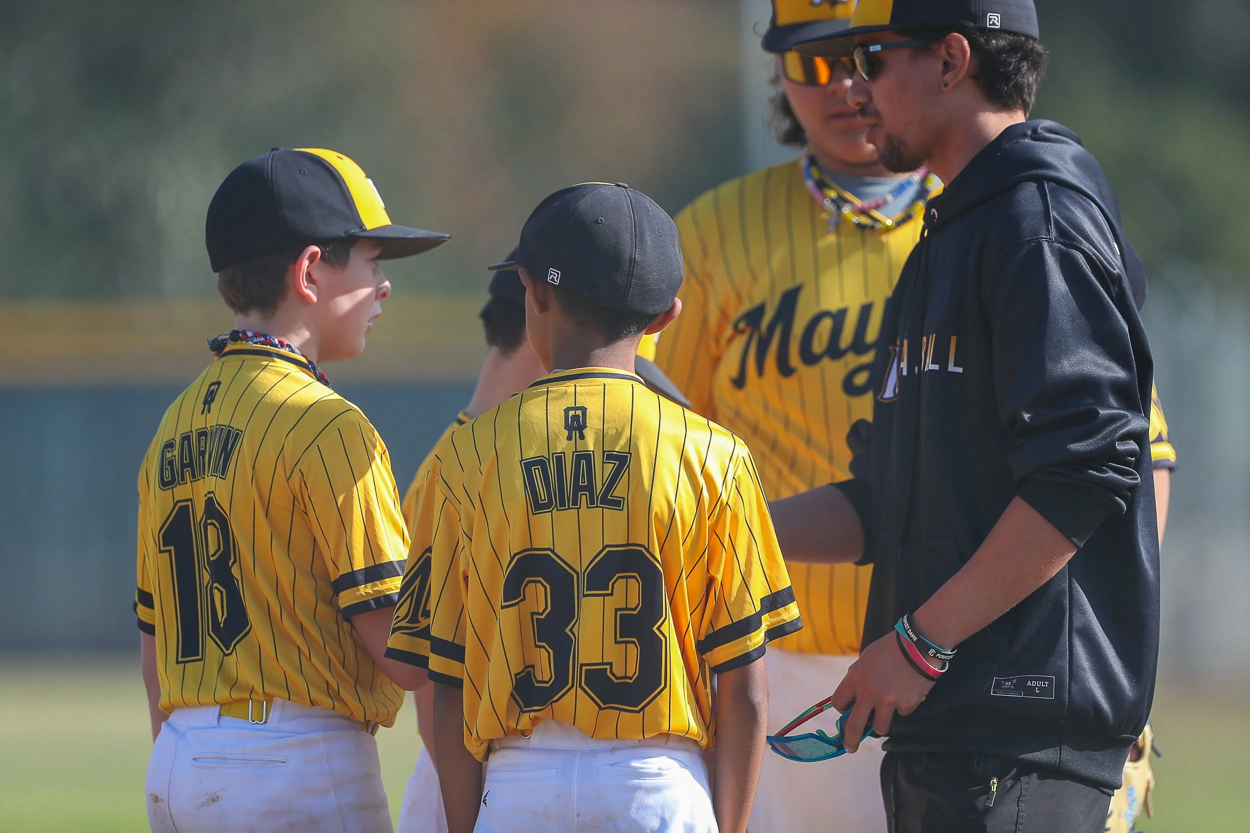 A youth baseball team in yellow striped jerseys with black lettering and numbers, including players Gairn and Diaz, standing together on a field while a coach, dressed in a dark jacket, talks to them during daylight.