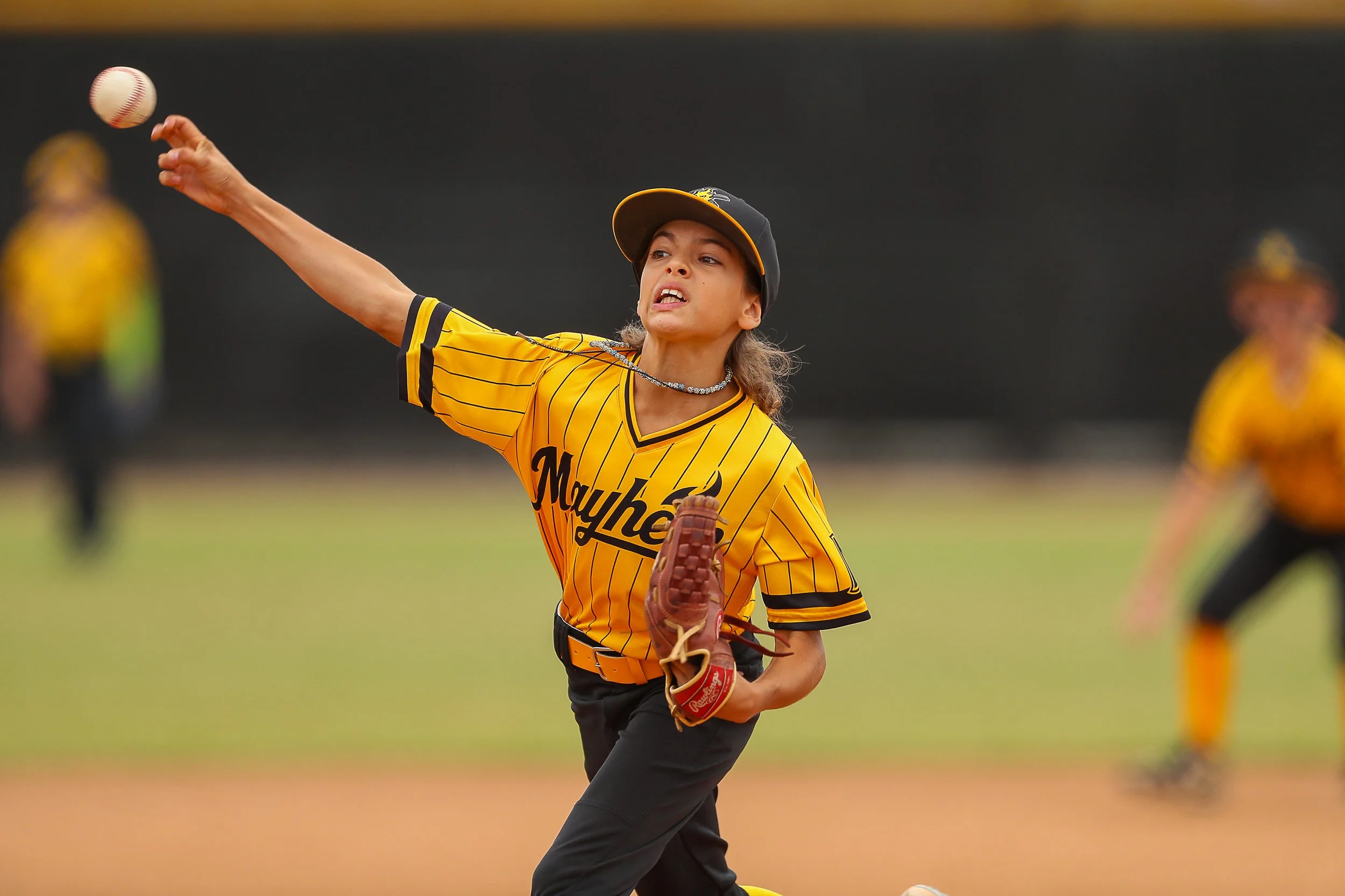 A young girl in a yellow and black baseball uniform throws a baseball on a field, with other players in matching uniforms in the background.