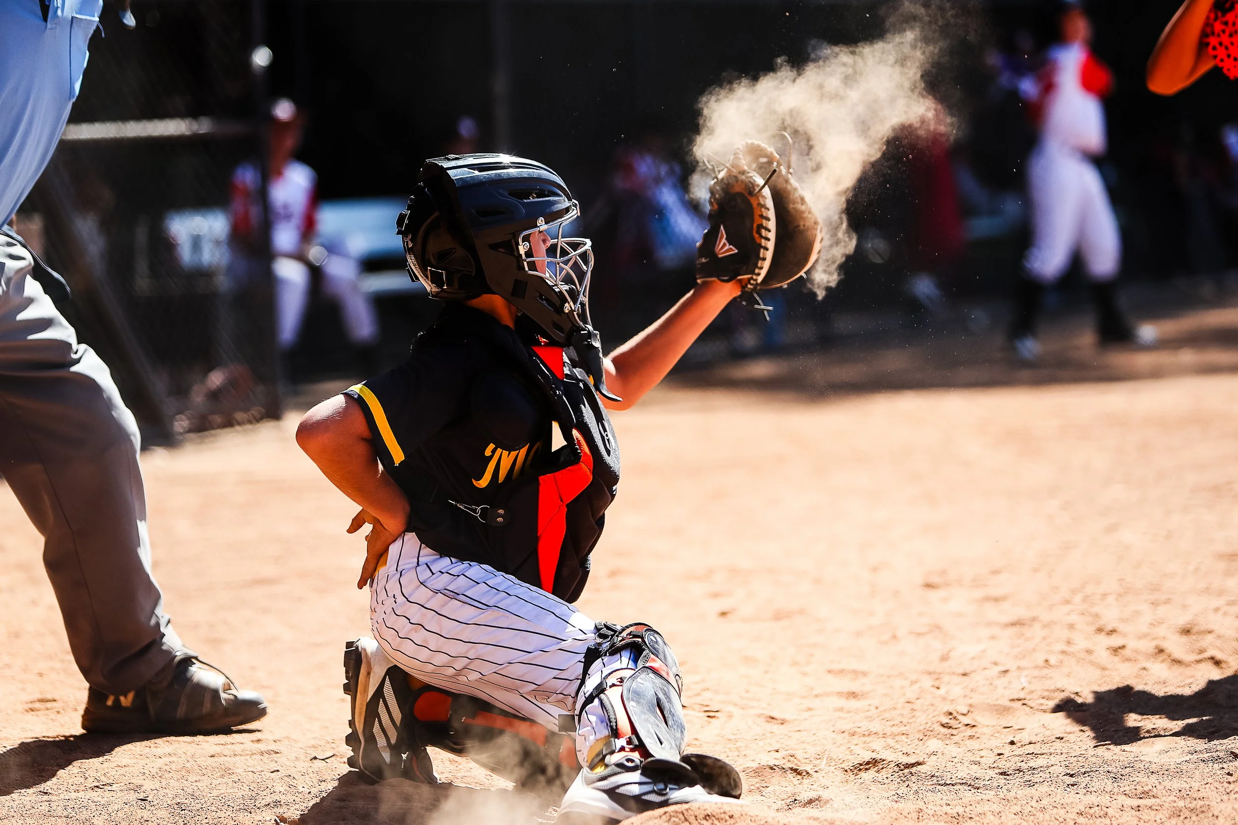 A young baseball catcher in black and white pinstriped uniform is kneeling on the dirt, catching a ball with glove, Dust is rising around him. A coach or umpire is standing nearby, and players and spectators are visible in the background.