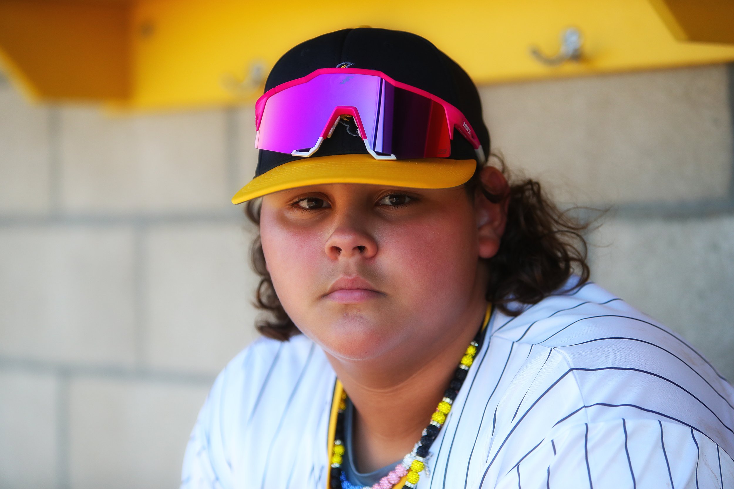 A young person wearing a black and yellow baseball cap with a pink sports sunglasses visor on top, a white striped sports jersey, and a colorful beaded necklace, sitting against a wall.