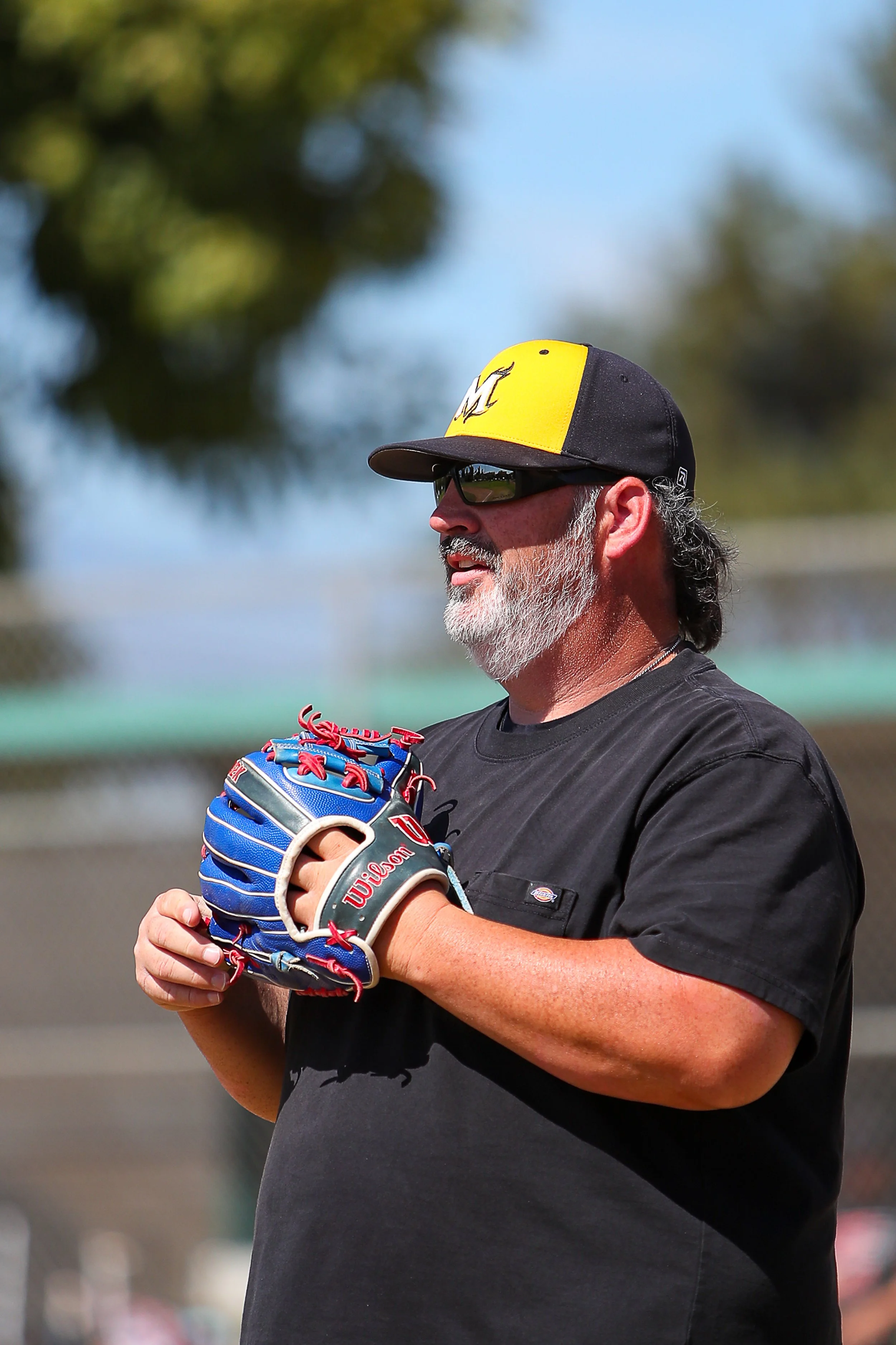 Coach Bill Rogers sitting down smiling wearing a black and yellow hat with an M logo on it.