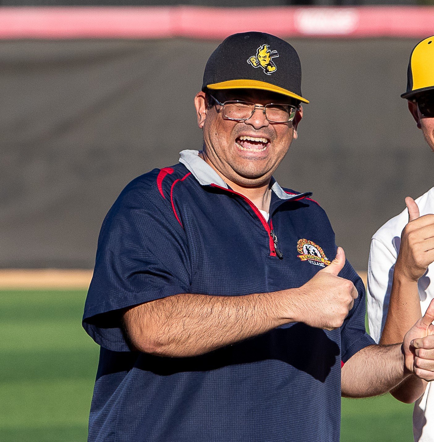 Coach Richard giving a thumbs-up gesture, wearing glasses, a dark blue sports shirt with red accents, and a black hat with a yellow brim and a logo.