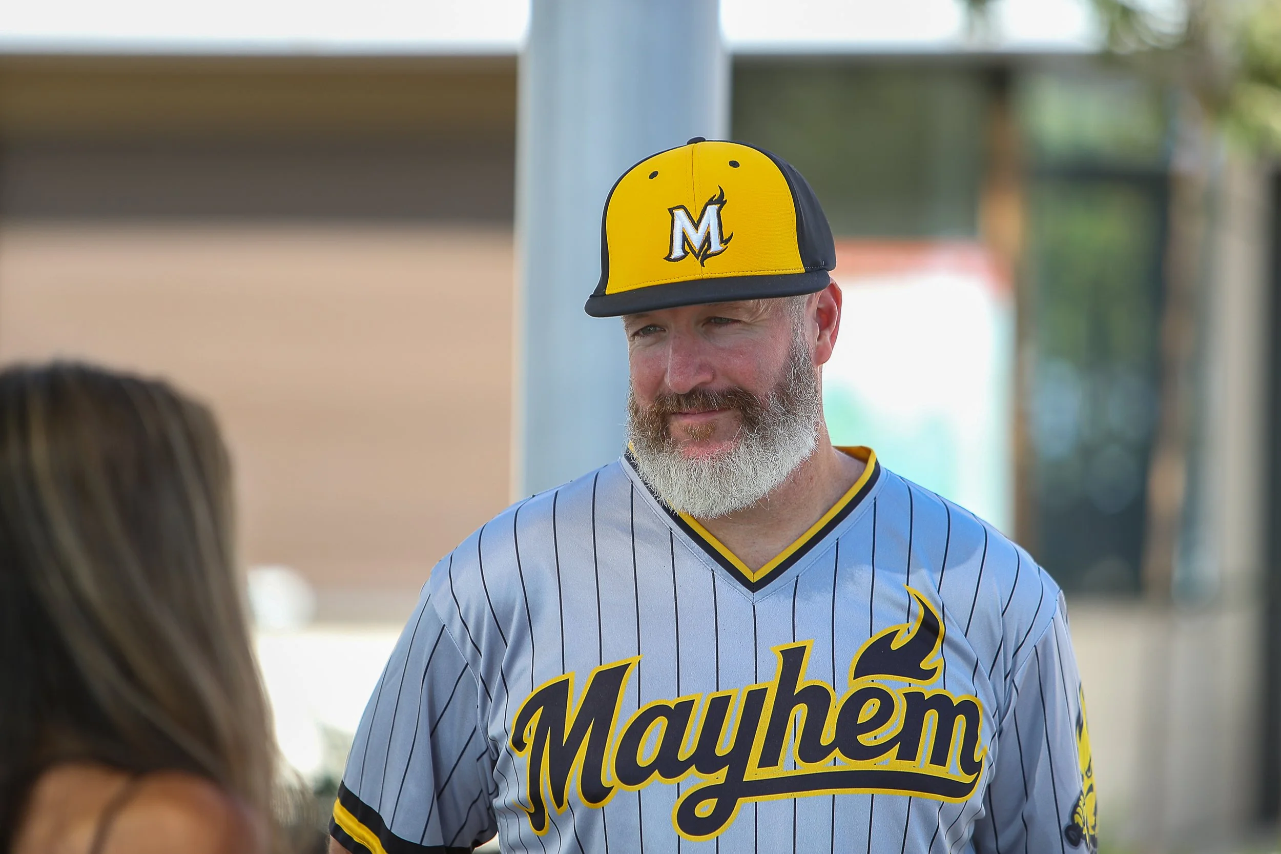 A man wearing a Mayhem baseball jersey and a yellow and black cap with an 'M' logo, standing outdoors and talking to a woman with long hair.
