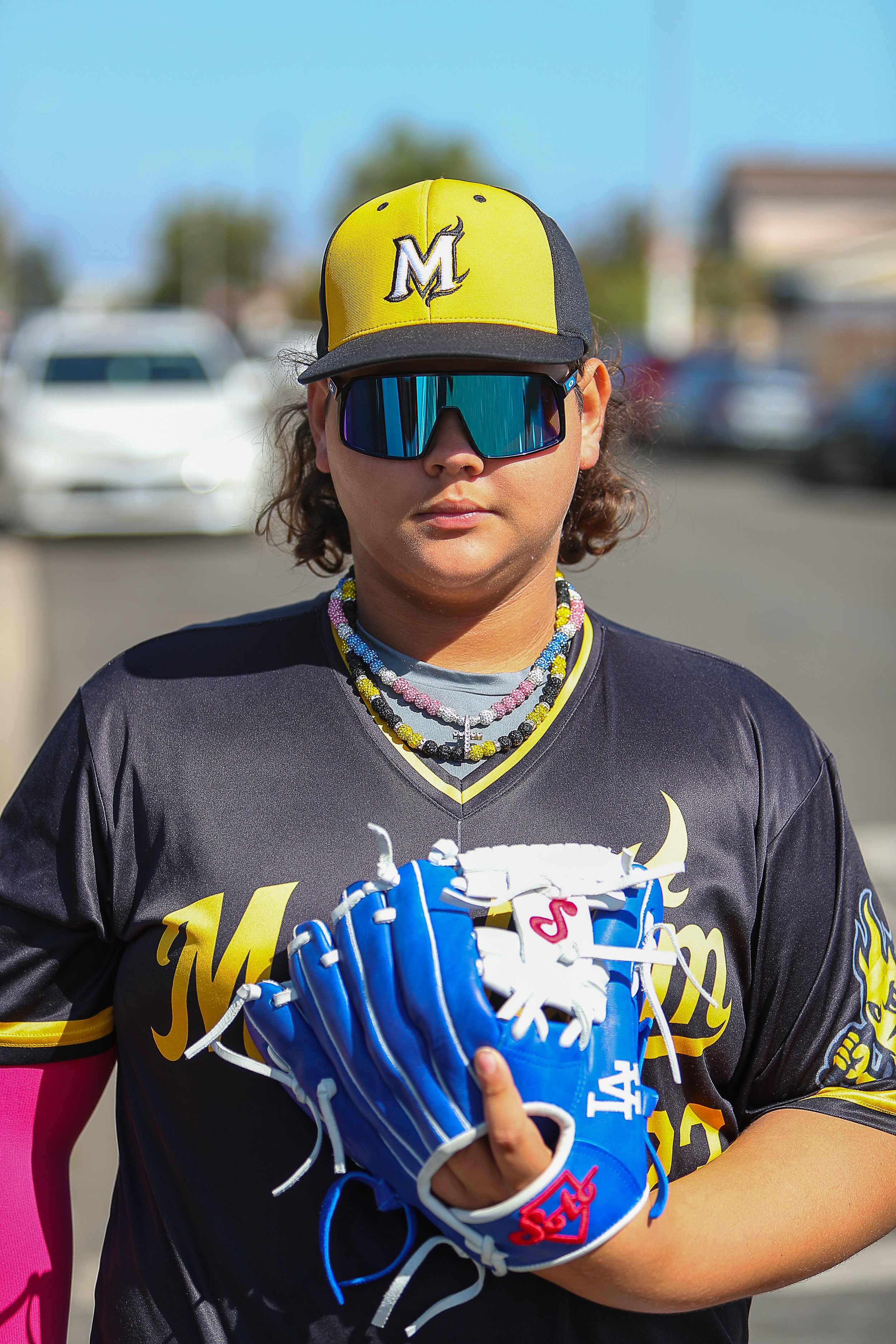 A young woman wearing a black and yellow baseball jersey, sports sunglasses, a yellow and black cap, and multiple colorful beaded necklaces. She holds a blue baseball glove in her left hand.