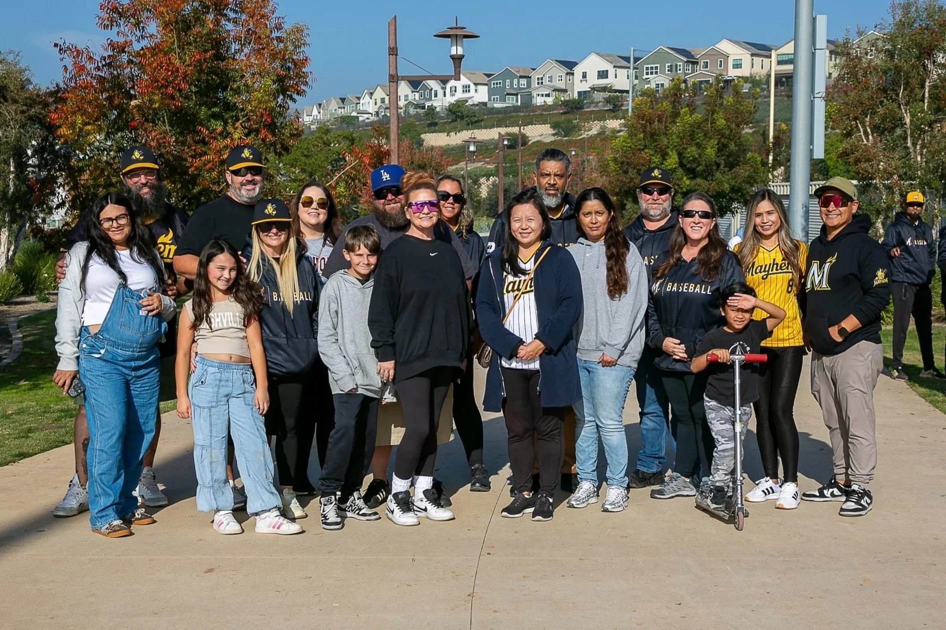 A group of youth baseball fans comprised of adults and children outside on a sunny day, standing on a sidewalk with trees and houses in the background.