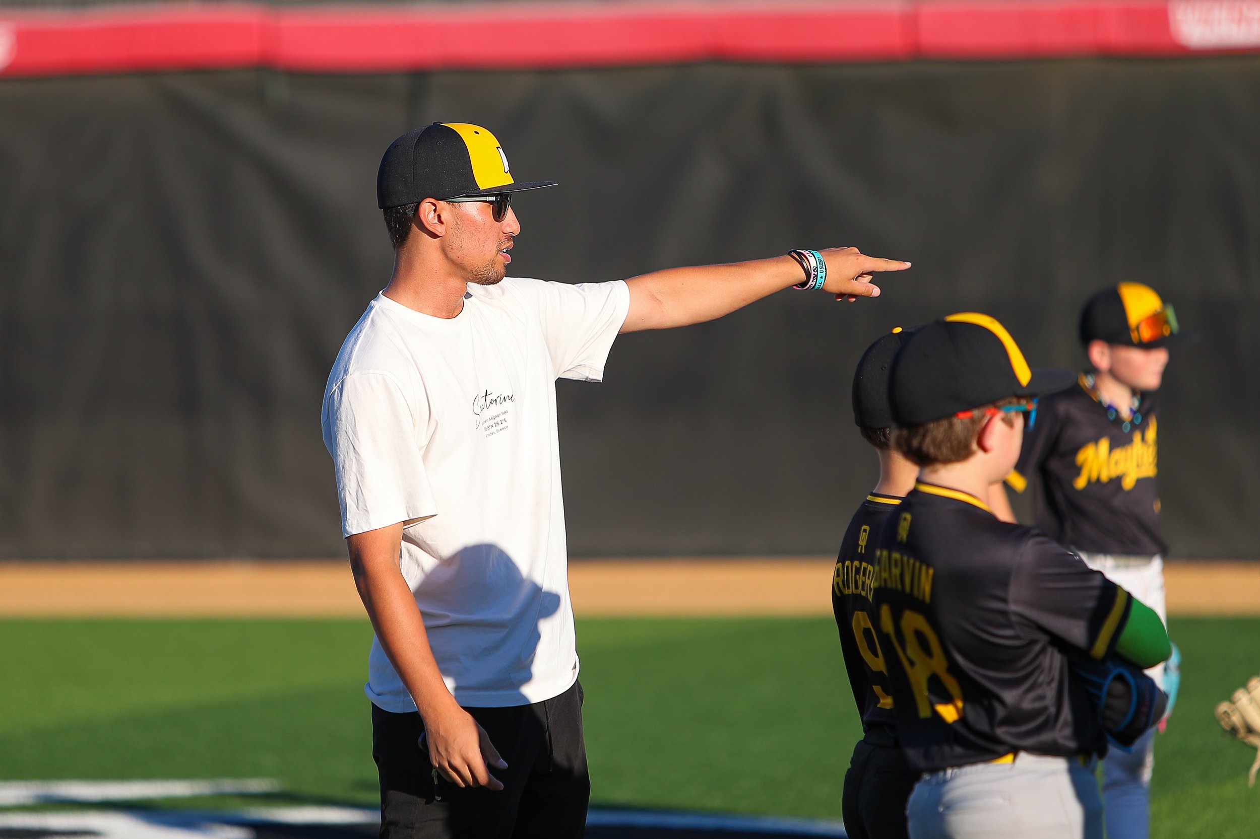 A baseball coach wearing a white T-shirt, black cap with yellow emblem, and sunglasses, is pointing while talking to young baseball players on a field during a game or practice.