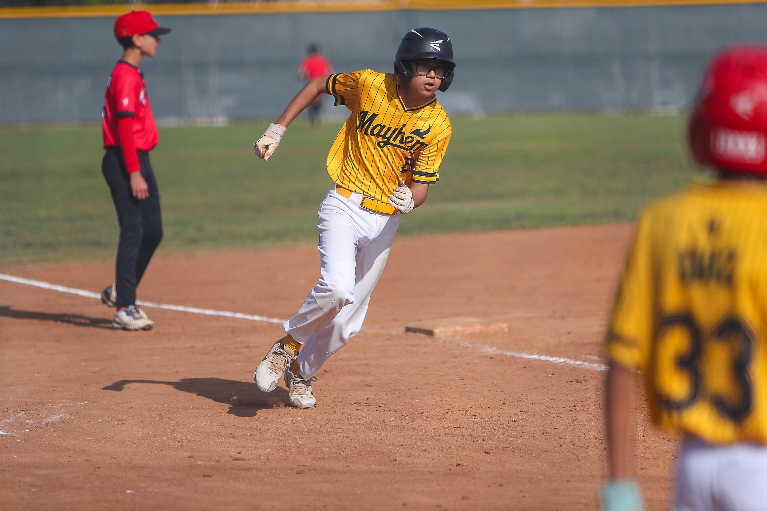 A young baseball player in a yellow and black uniform running on the field during a game, wearing a helmet and glasses.