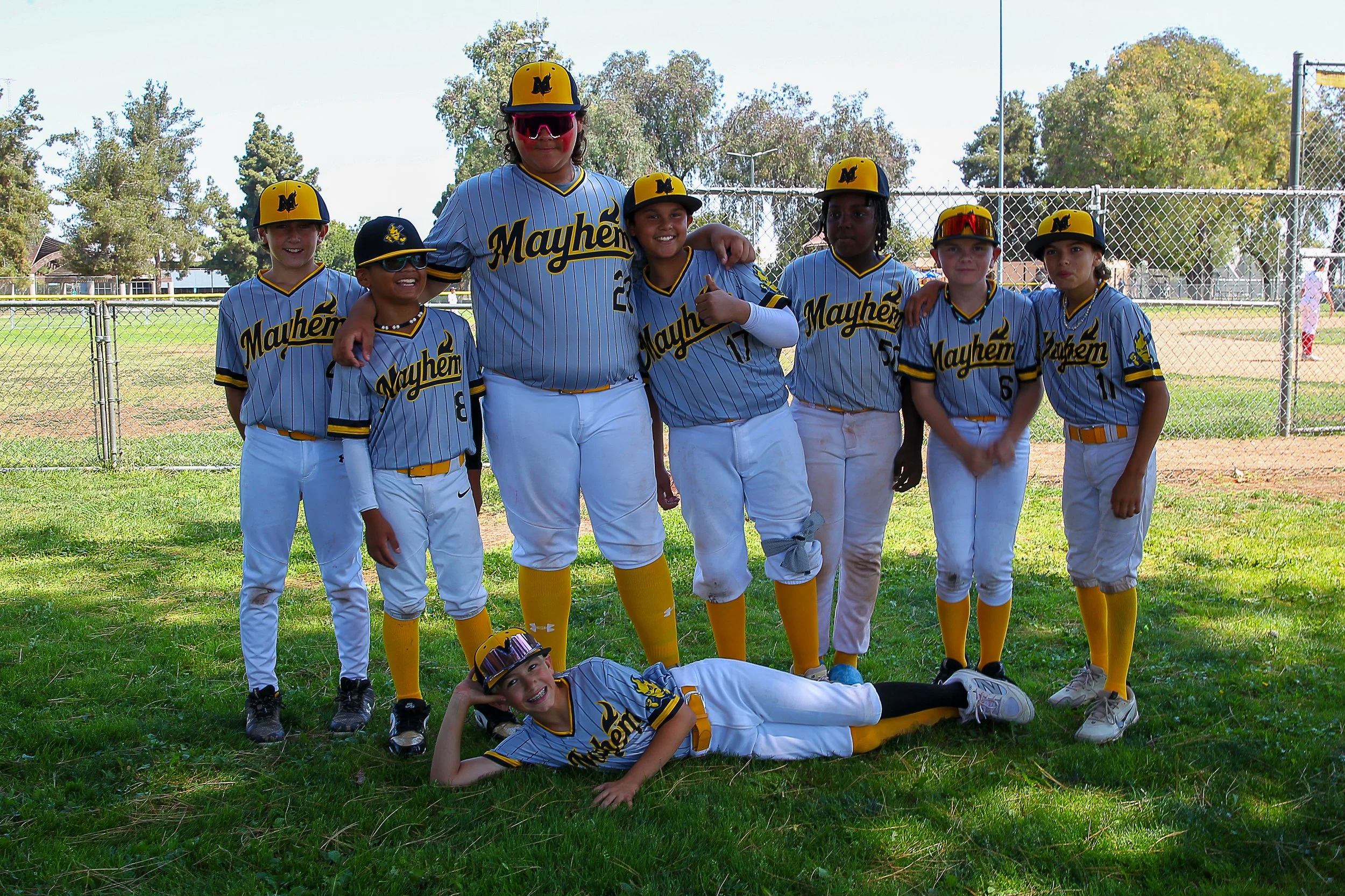 Youth baseball team in matching uniforms, posing on a grassy field with a chain-link fence and trees in the background.