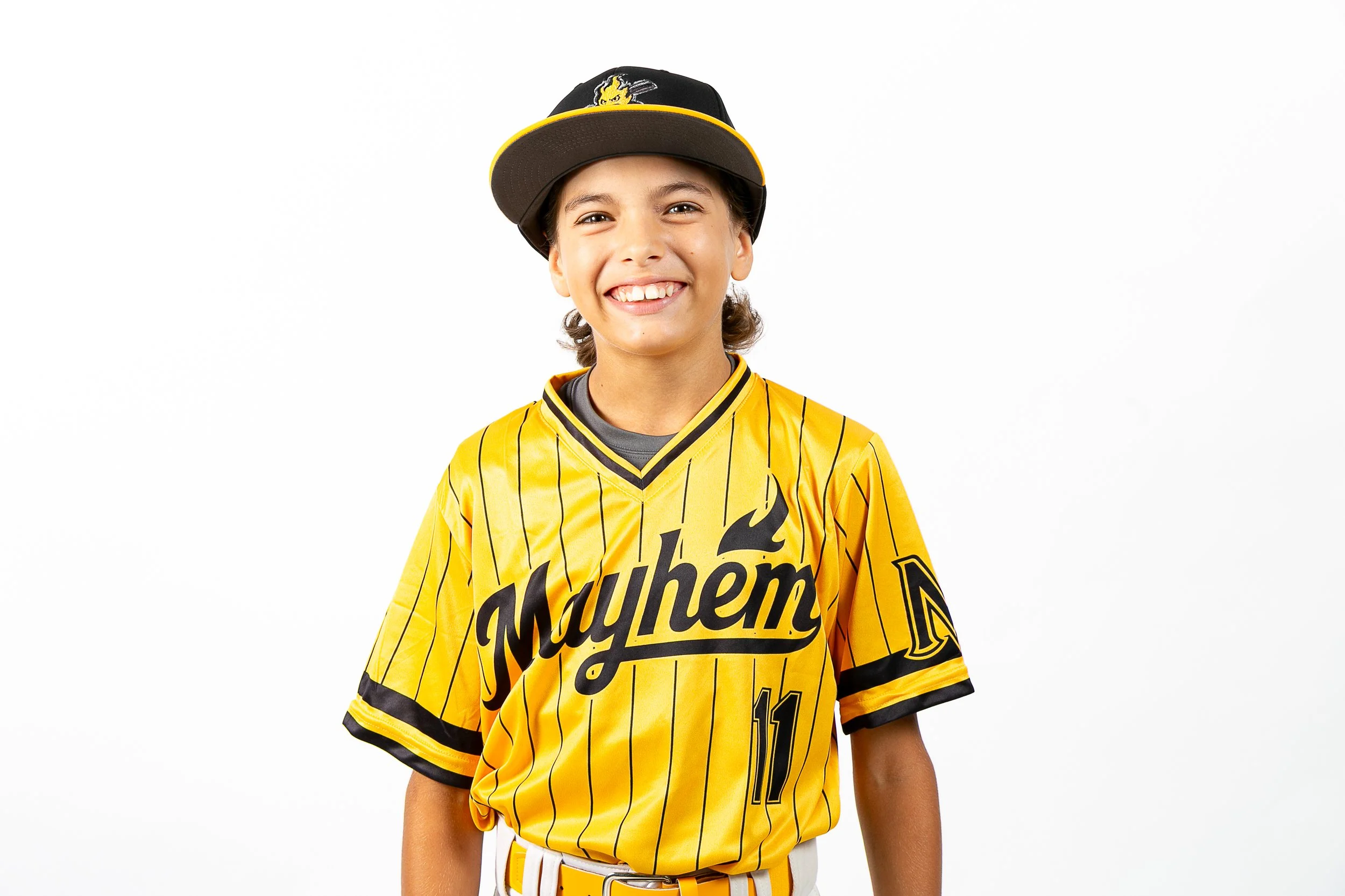 Young boy smiling, wearing a yellow baseball jersey with black pinstripes, a black and yellow cap tilted to the side, and a baseball belt.