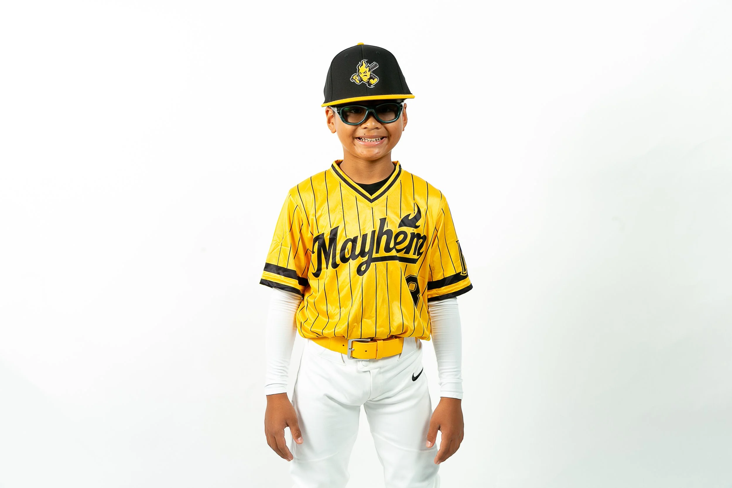 Young boy wearing a yellow baseball jersey with 'Mayhem' written on it, white pants, black cap with a small flame logo, sunglasses, and a yellow belt, standing against a plain white background.