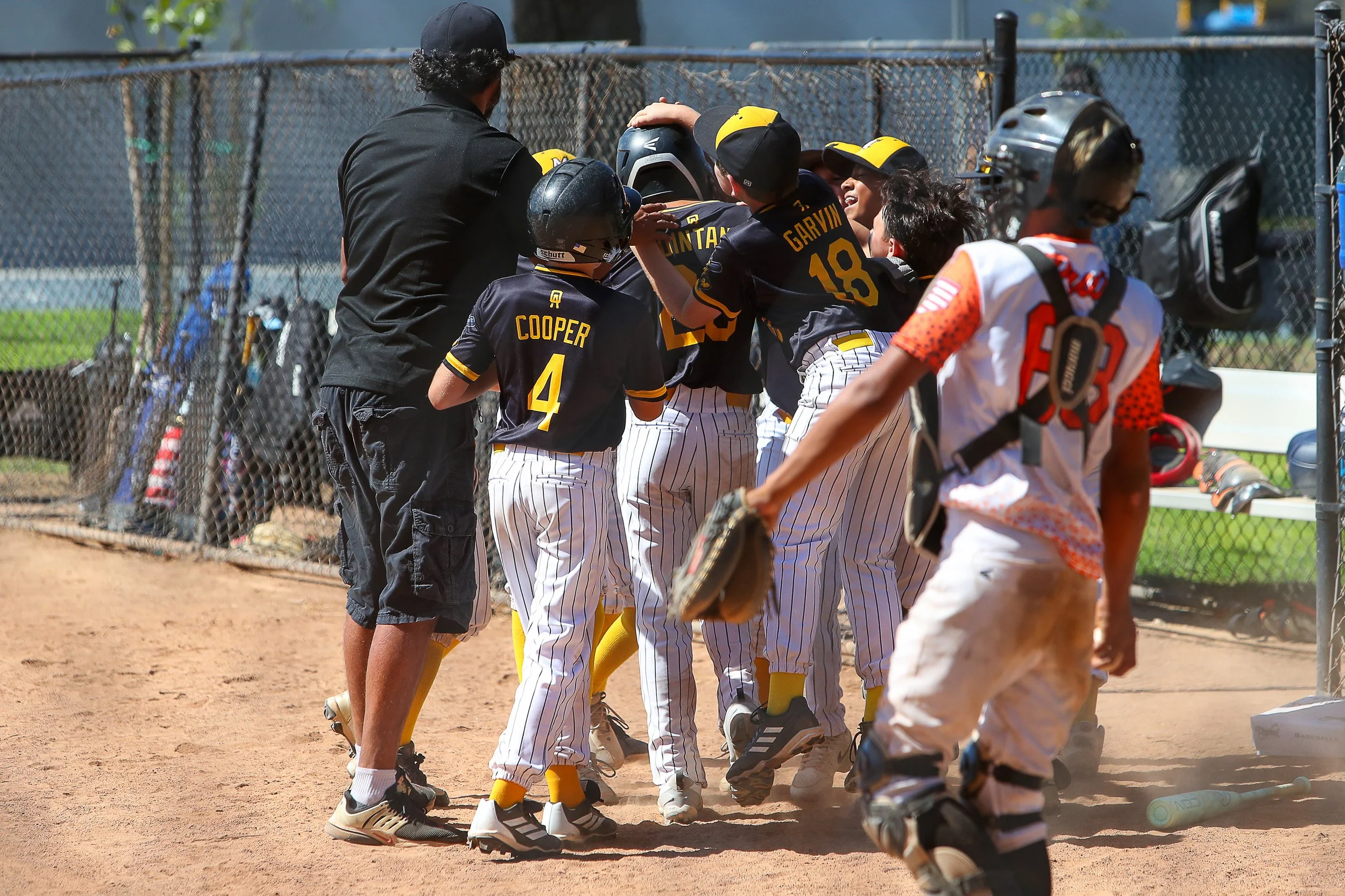 Youth baseball team celebrating on the field with coach, kids wearing black and white pinstripe uniforms and protective gear, some with helmets, near the dugout with equipment.