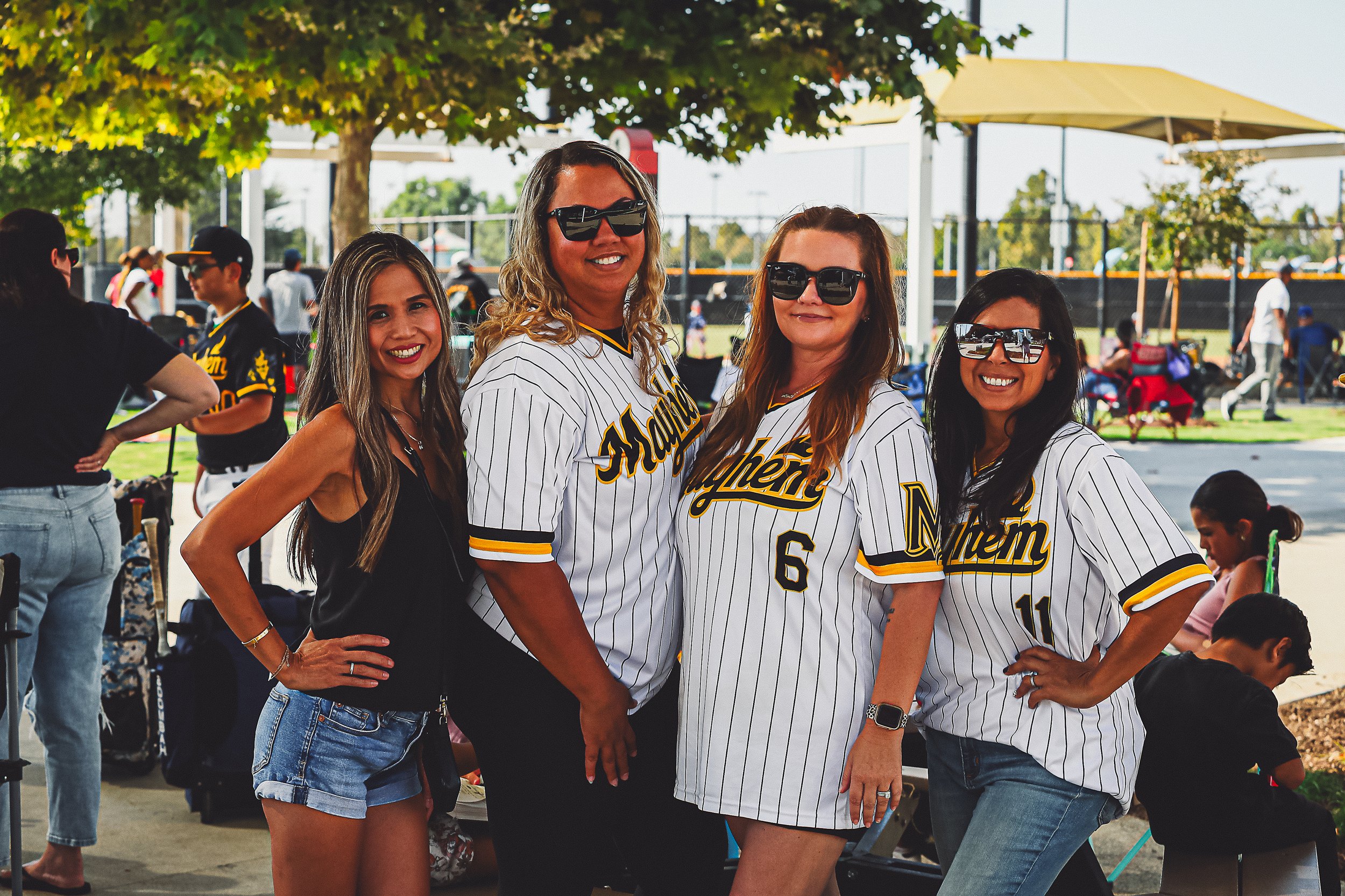 Four women at a baseball game, some wearing sunglasses and jerseys with 'Mayhem' on them, standing outdoors on a sunny day, with a baseball field and other spectators in the background.