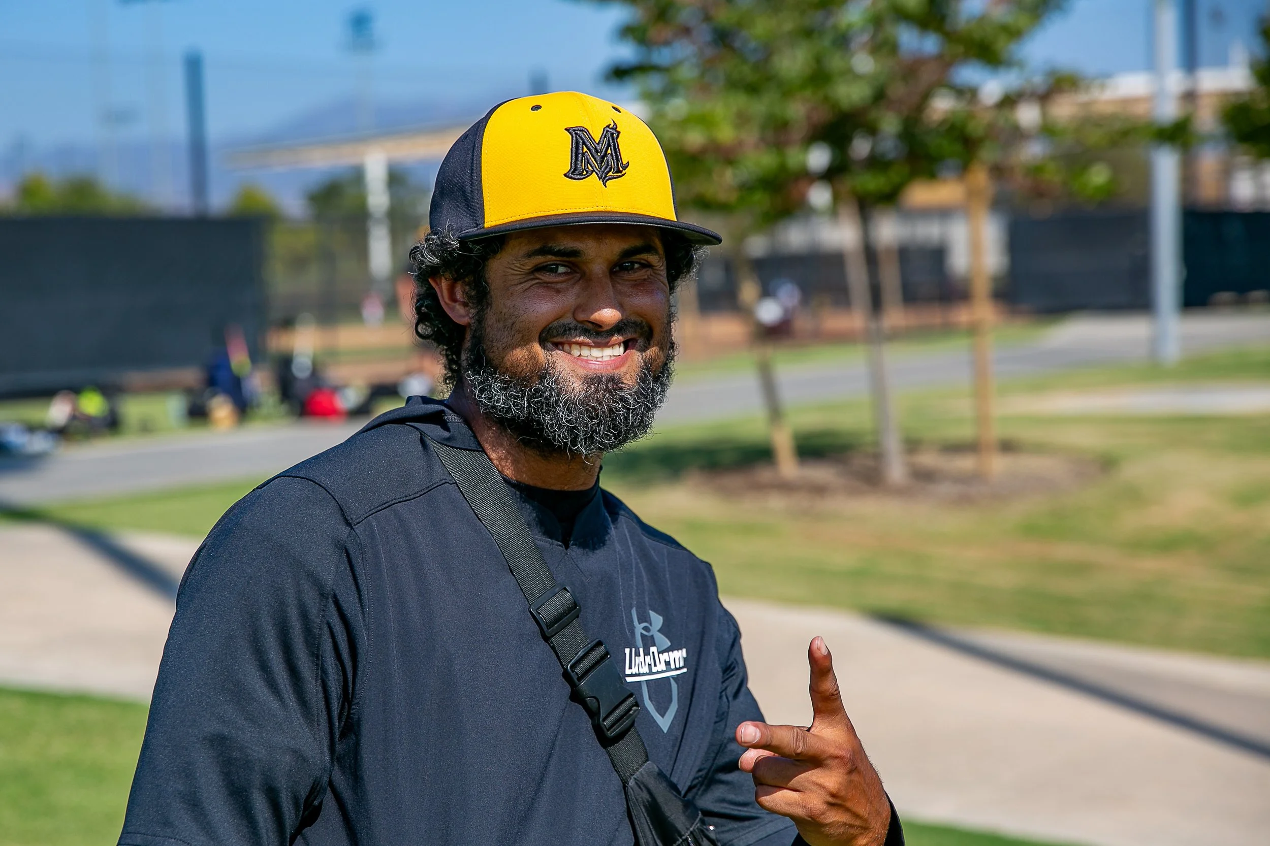 Coach Robert wearing a yellow and black Mayhem cap and a dark sports shirt, pointing with his index finger outdoors in a park.