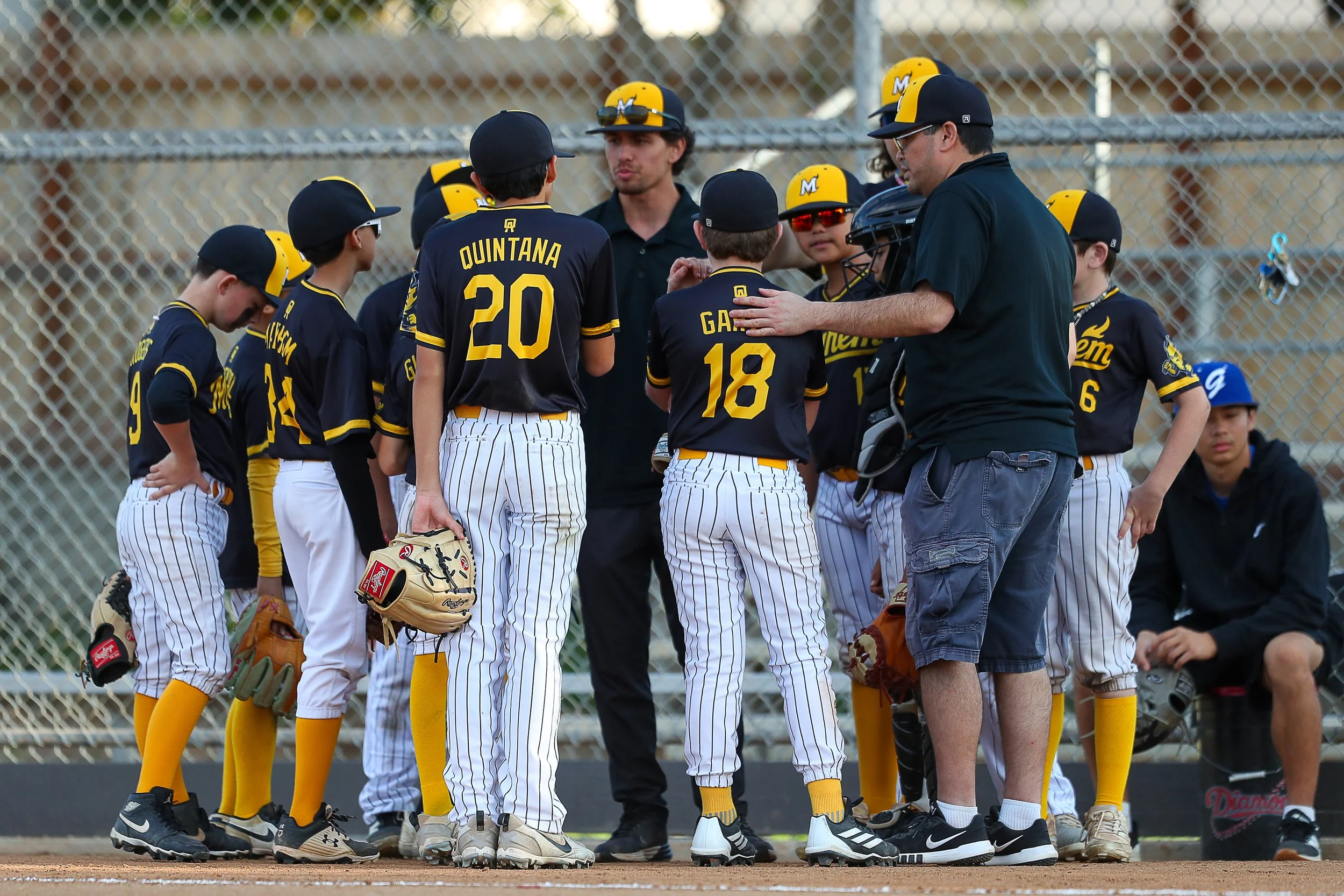 Young baseball players in black and yellow uniforms gather around their coach on a baseball field.