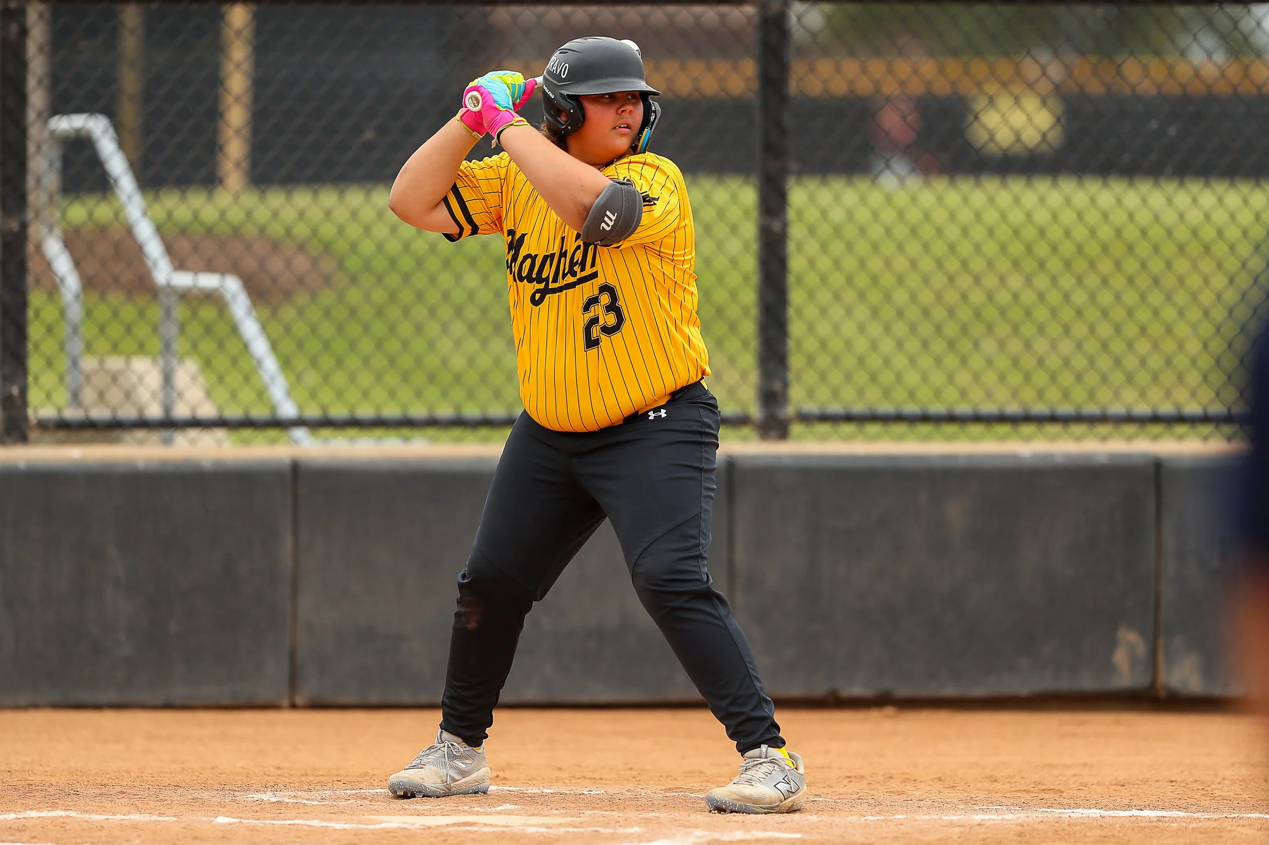 A young female softball player wearing a yellow jersey with black pinstripes, black pants, a helmet, and pink batting gloves stands at home plate ready to bat, on a dirt field with a chain-link backstop and a grassy area in the background.
