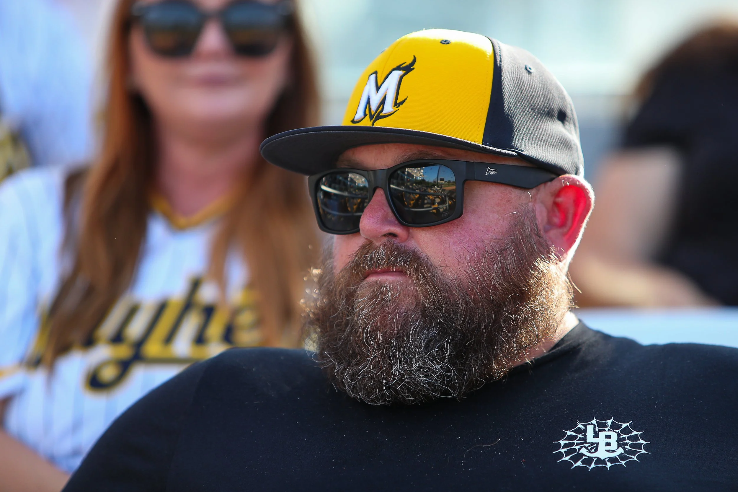 A man with a gray beard and red cheeks wearing black sunglasses and a yellow and gray cap with a large letter M. He is also wearing a black shirt with a logo in the shape of a spider web. In the background, a woman with long red hair, sunglasses, and a baseball jersey is visible but out of focus.