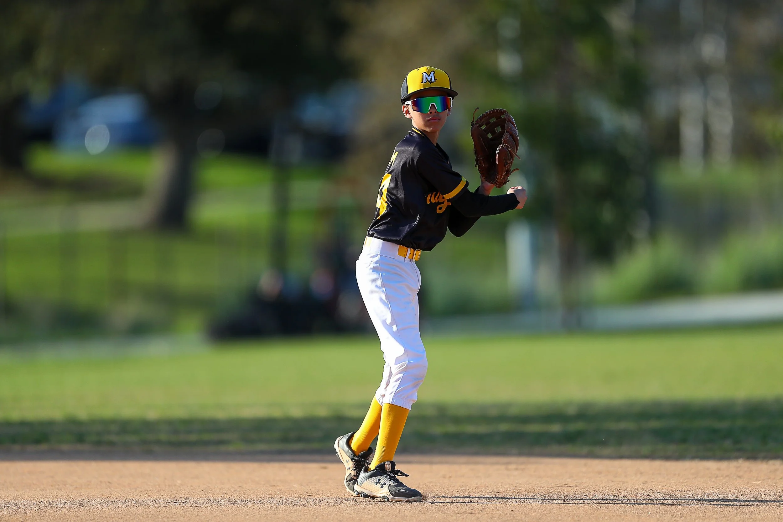 Young baseball player on field, wearing a yellow baseball hat, sunglasses, black and yellow jersey, white pants, yellow socks, and holding a baseball glove, in ready position.