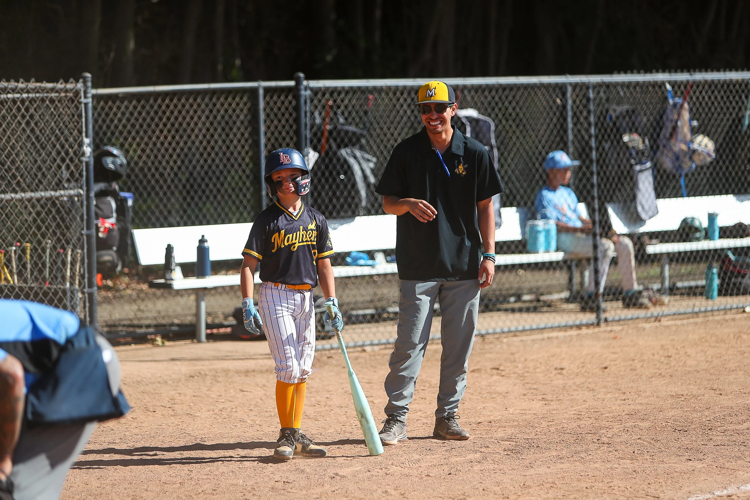 A young baseball player in uniform standing on a baseball field holding a bat, with a man smiling and talking to him. Other players and spectators are in the background.