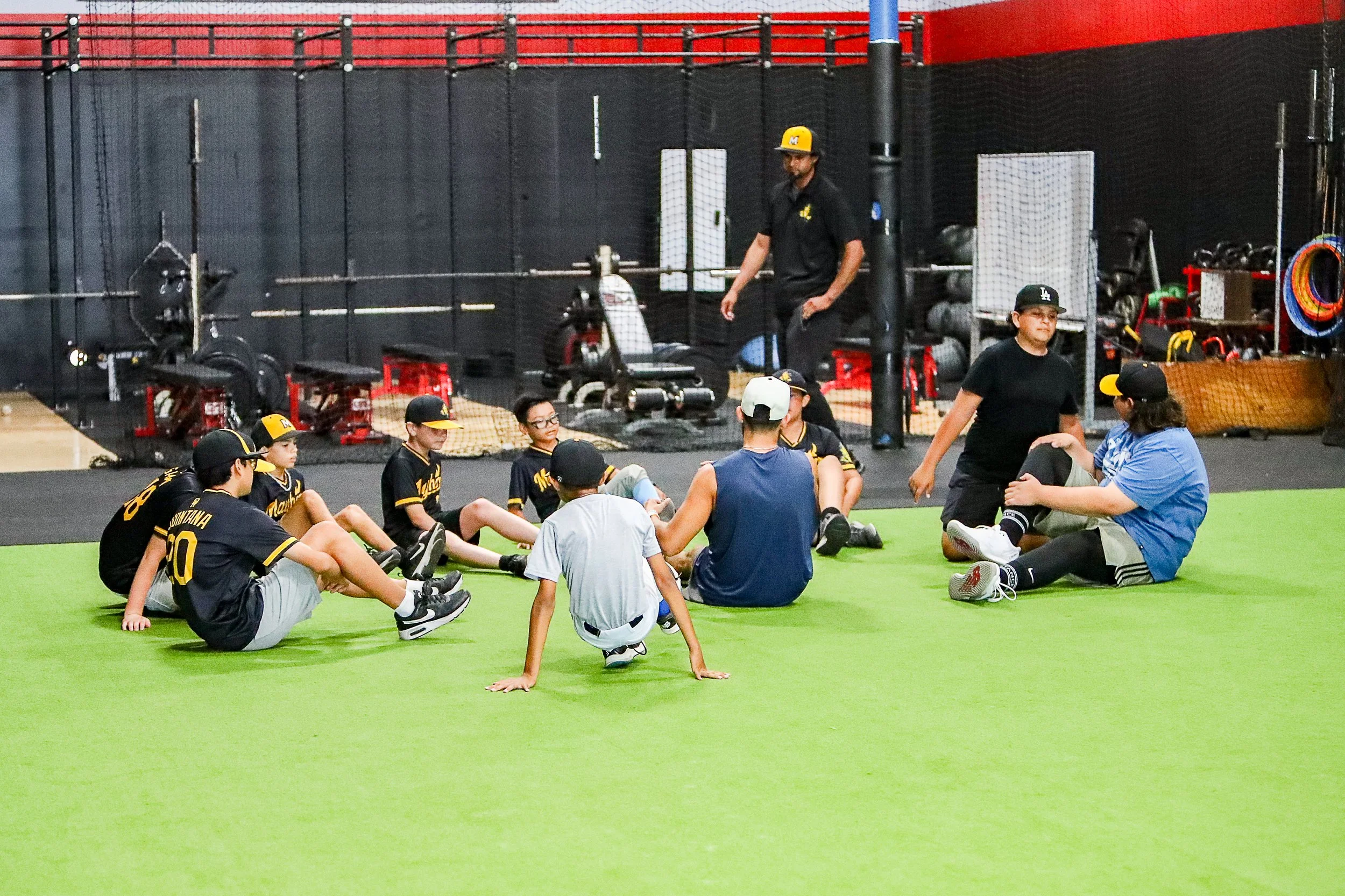 A group of young baseball players and coaches sitting and talking on an indoor turf field doing speed and agility workouts, with equipment storage and gym equipment in the background.