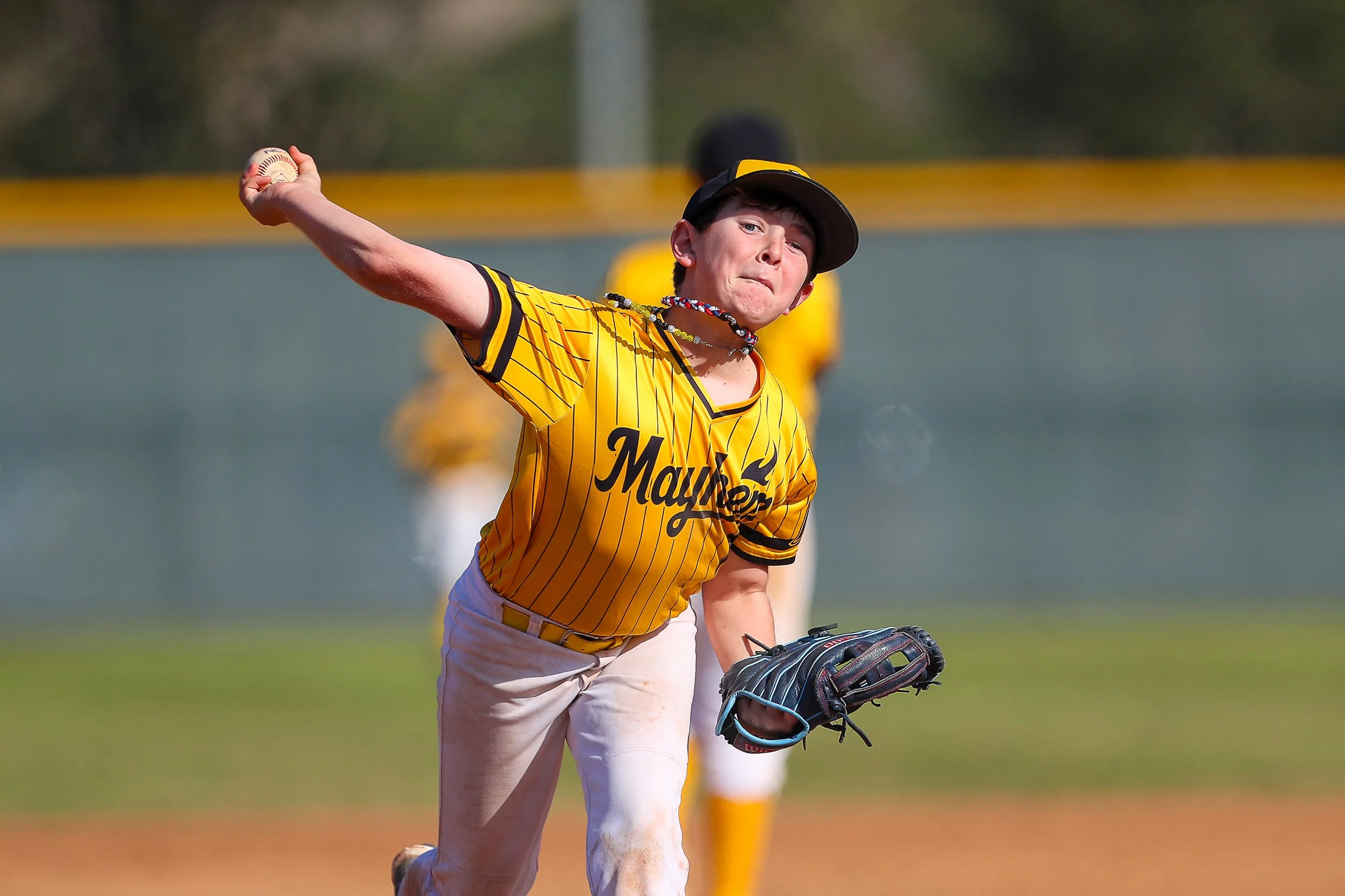 A young baseball player in a yellow and black jersey with the word 'Mayhem' on it, pitching a ball on a baseball field during daytime. The player is wearing a black hat and a necklace, with other players visible in the background.