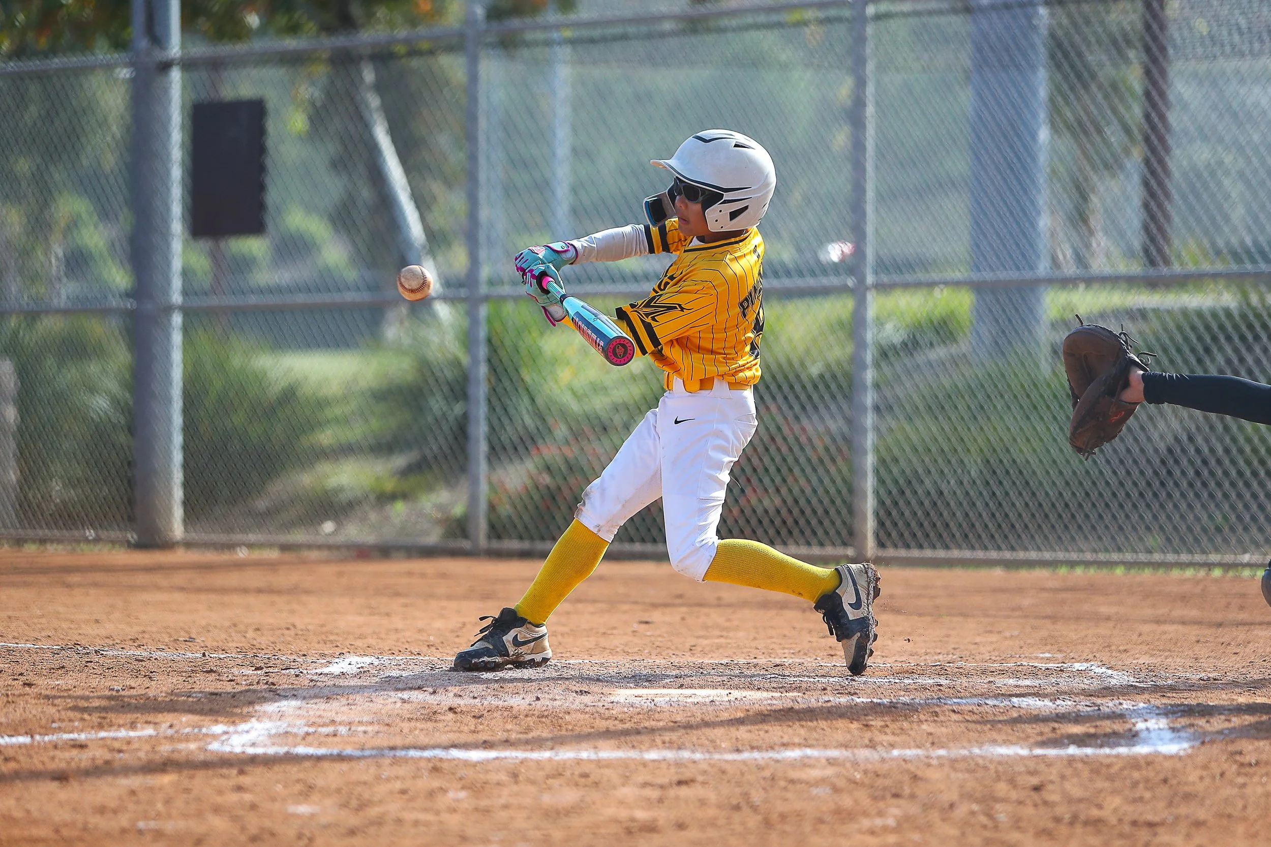 A young baseball player in a yellow jersey and white pants swings a bat during a game on a baseball field, with a chain-link fence and trees in the background.