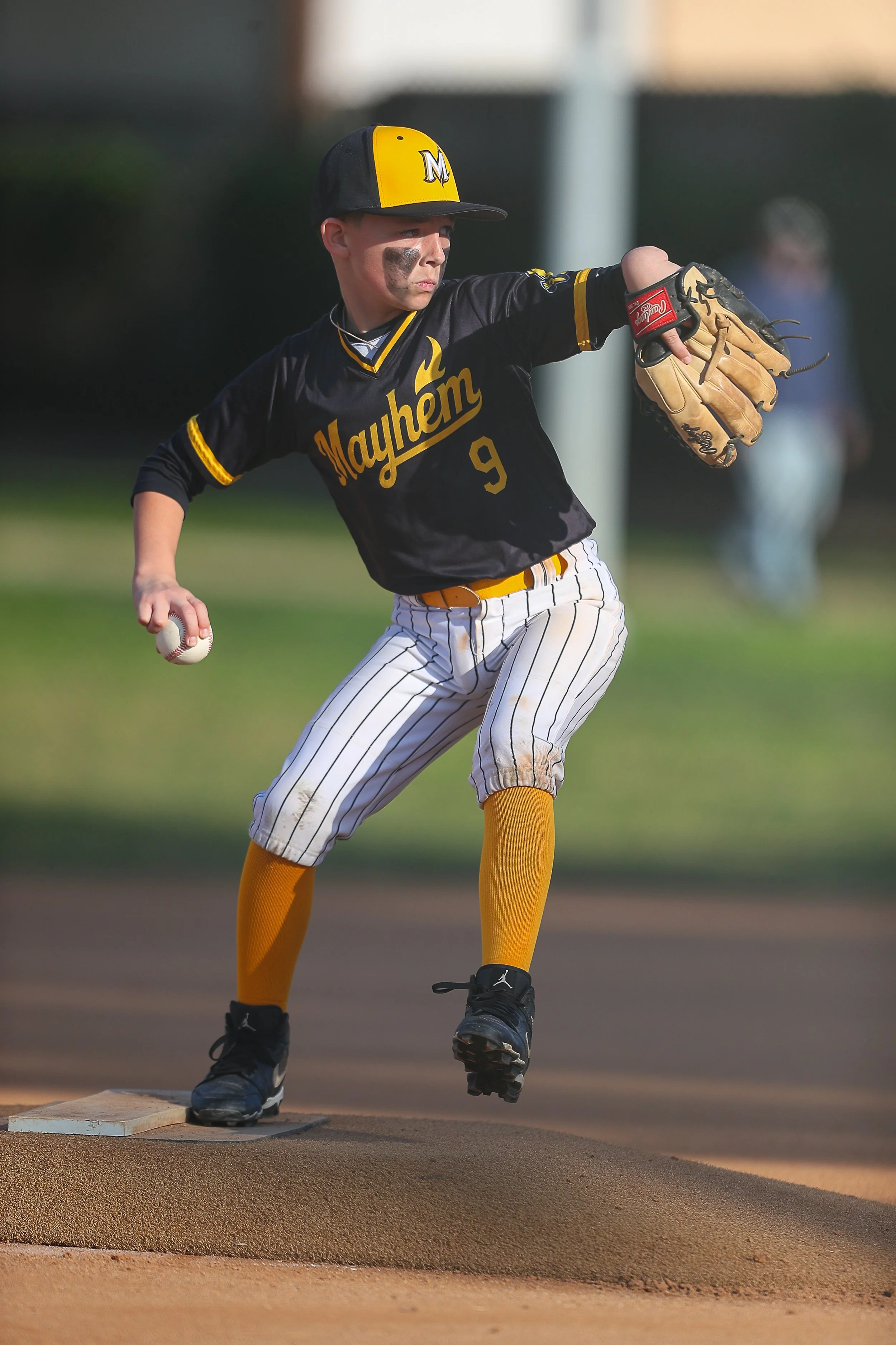 A young baseball player is pitching on a baseball field, wearing a black and yellow uniform with "Mayhem" written on it and the number 9, a black and yellow cap, yellow socks, and black sneakers, holding a baseball in his right hand and a glove on hi