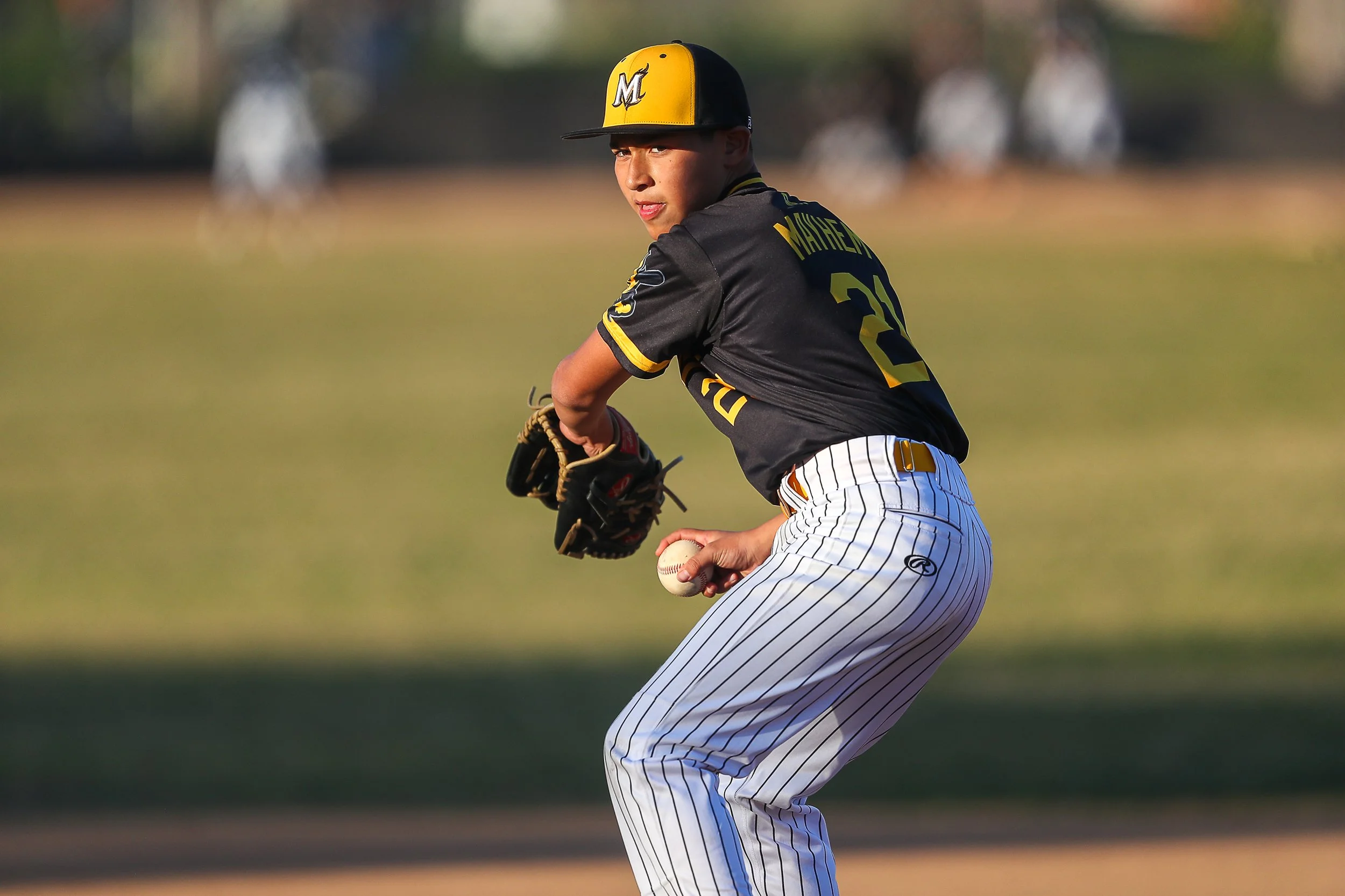 A young baseball player in a black and yellow uniform is preparing to throw a baseball, wearing a black glove, on a baseball field during daytime.