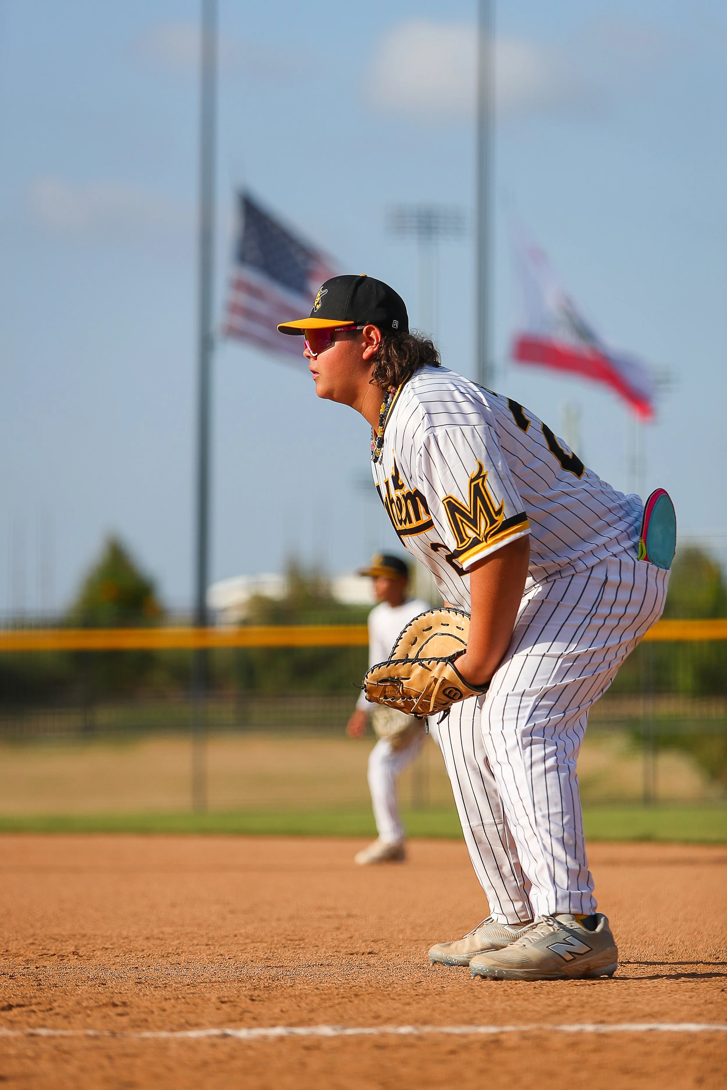A baseball player in a pinstriped white and black uniform crouching on the field, wearing sunglasses, a cap, and holding a baseball glove, with flags and another player in the background.