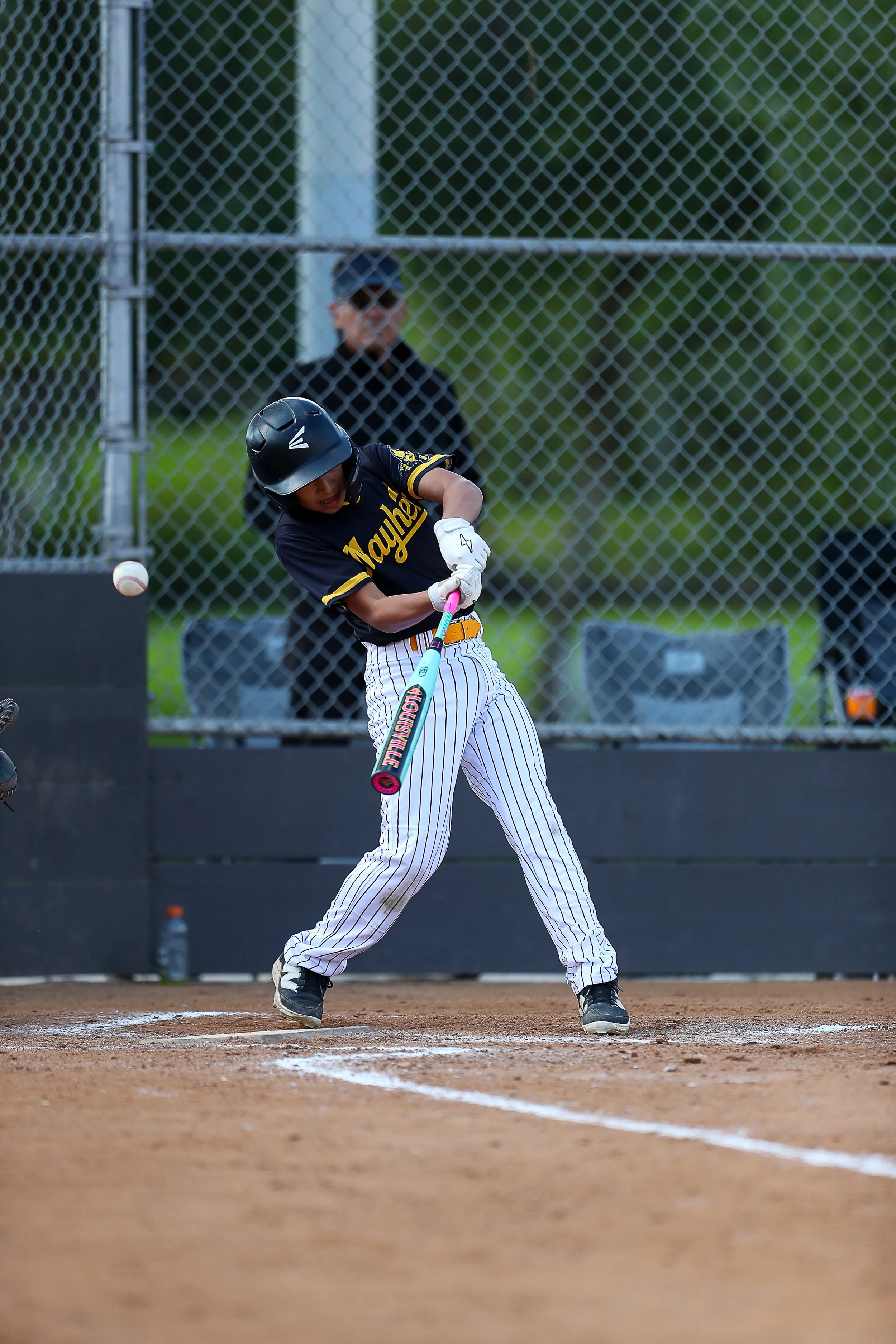 A young baseball player in a black uniform with white pinstripe pants is swinging a pink and blue Louisville baseball bat at a pitched ball on the field.