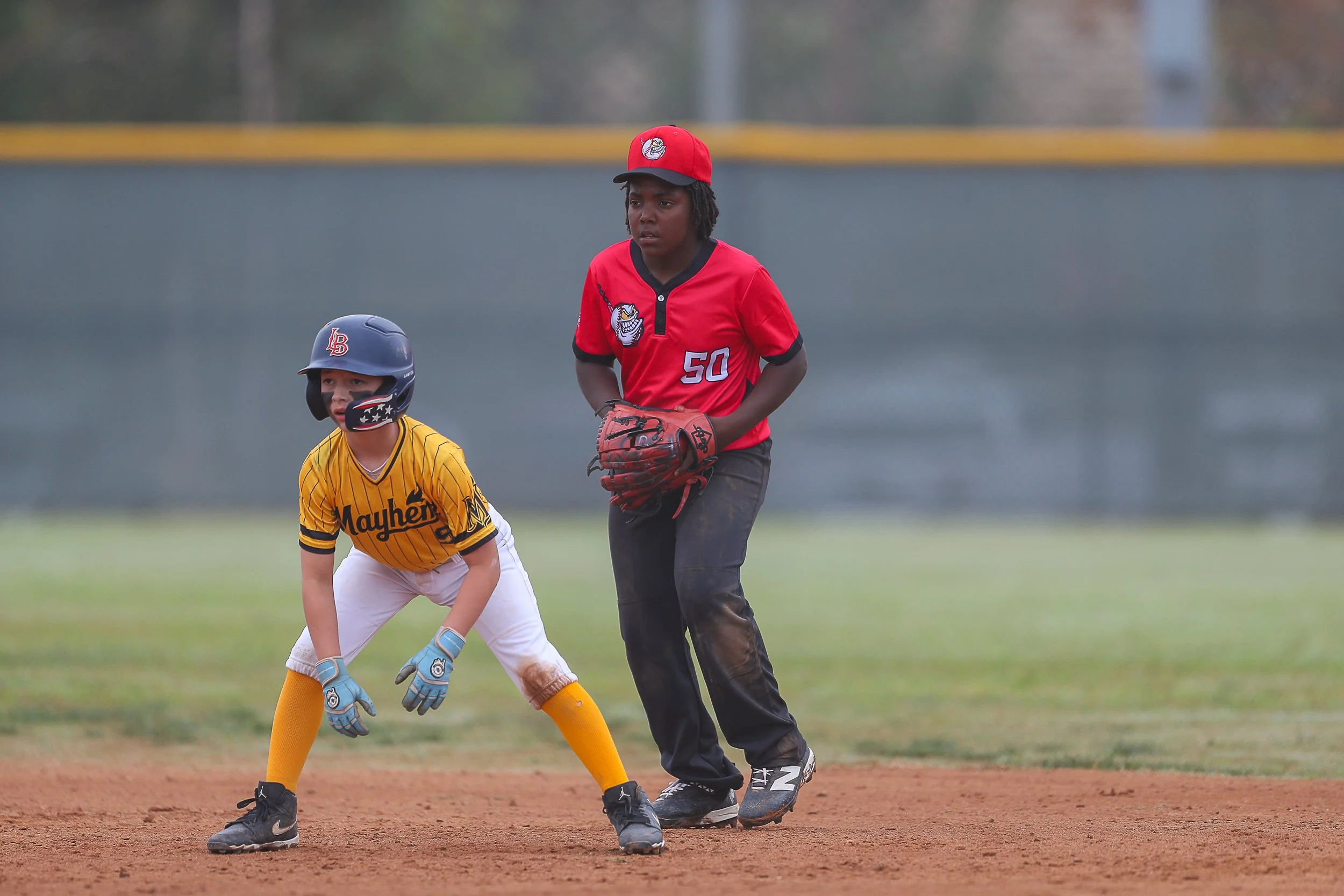 Two young baseball players on the field, one in a yellow jersey and helmet, the other in a red jersey and cap, preparing for a play.