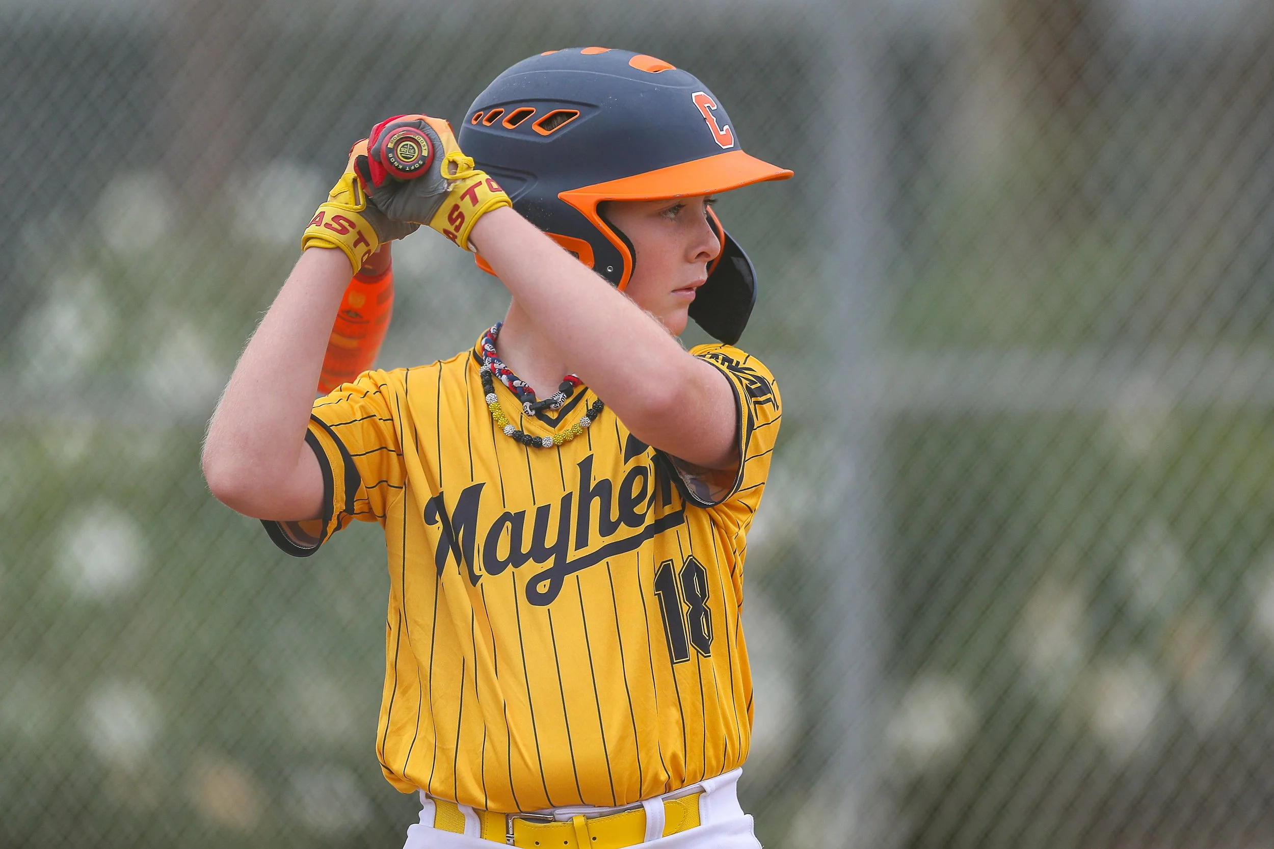A young female softball player in a yellow jersey with black pinstripes, number 18, preparing to bat. She is wearing a black and orange helmet, yellow batting gloves, and a necklace, standing on a field with a chain-link fence in the background.