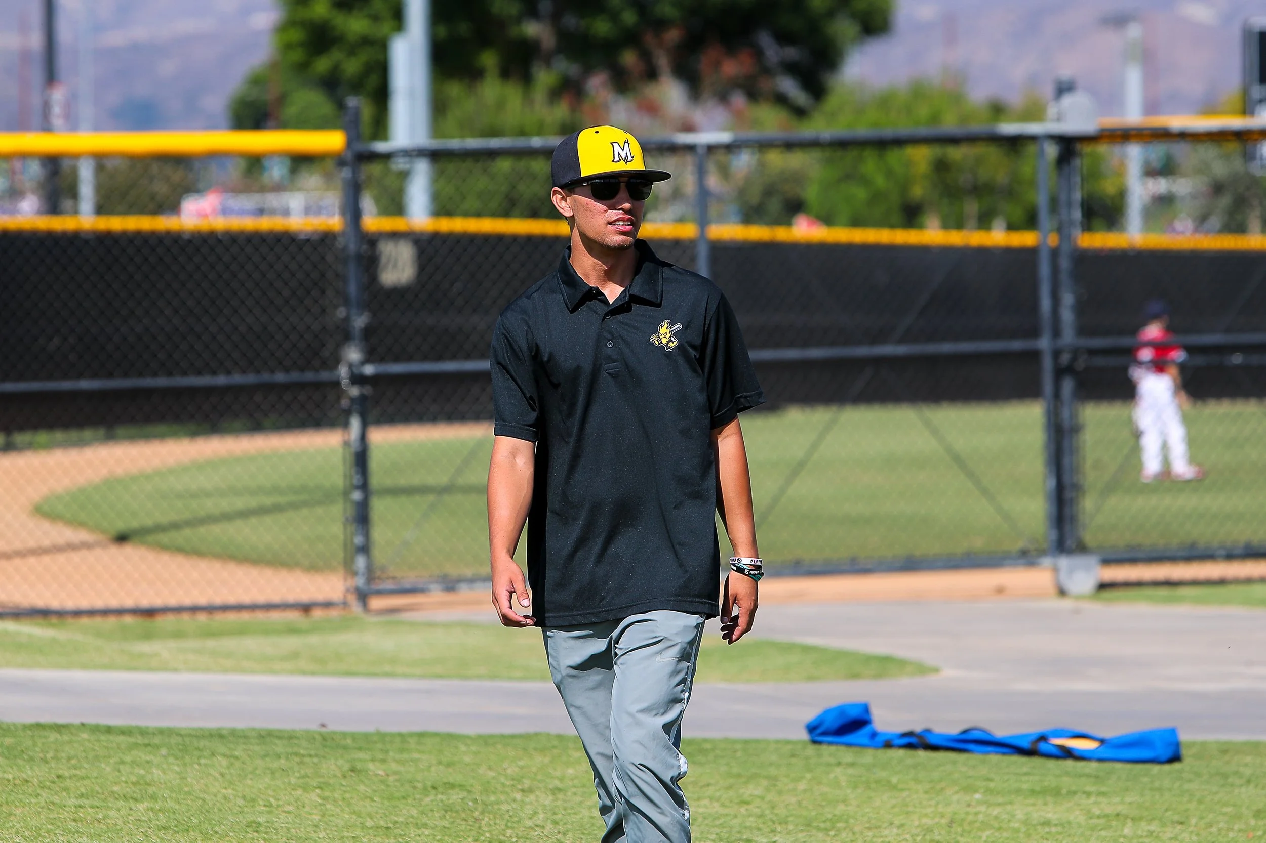 Coach Aaron wearing sunglasses, a black polo shirt with a sports team logo, gray pants, and a baseball cap with a yellow and black design walking on a baseball field.