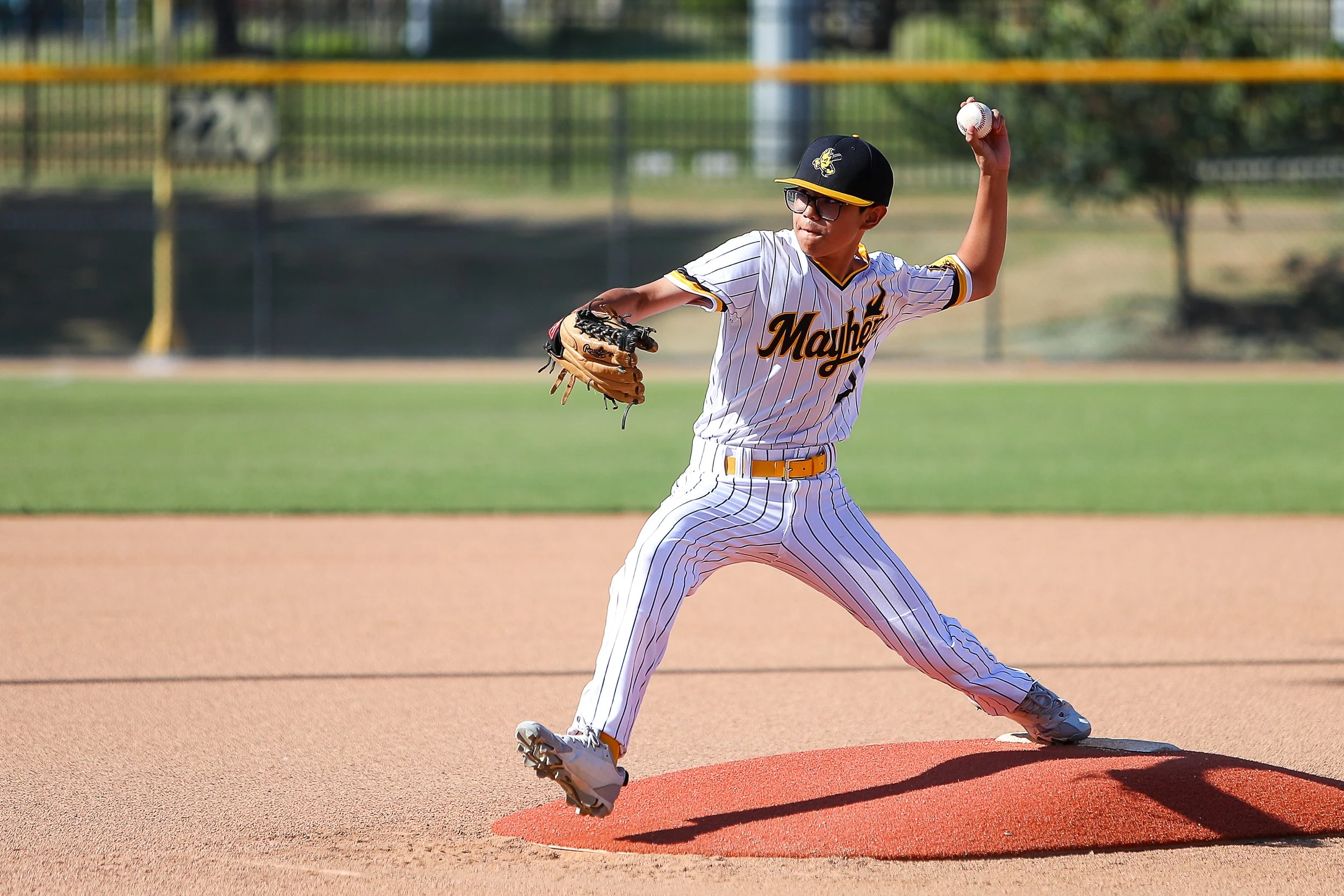 A young baseball player in a white pinstripe uniform with 'Mayhem' written on the front, wearing a black cap, sunglasses, and a glove, preparing to throw a baseball on a baseball field during daytime.