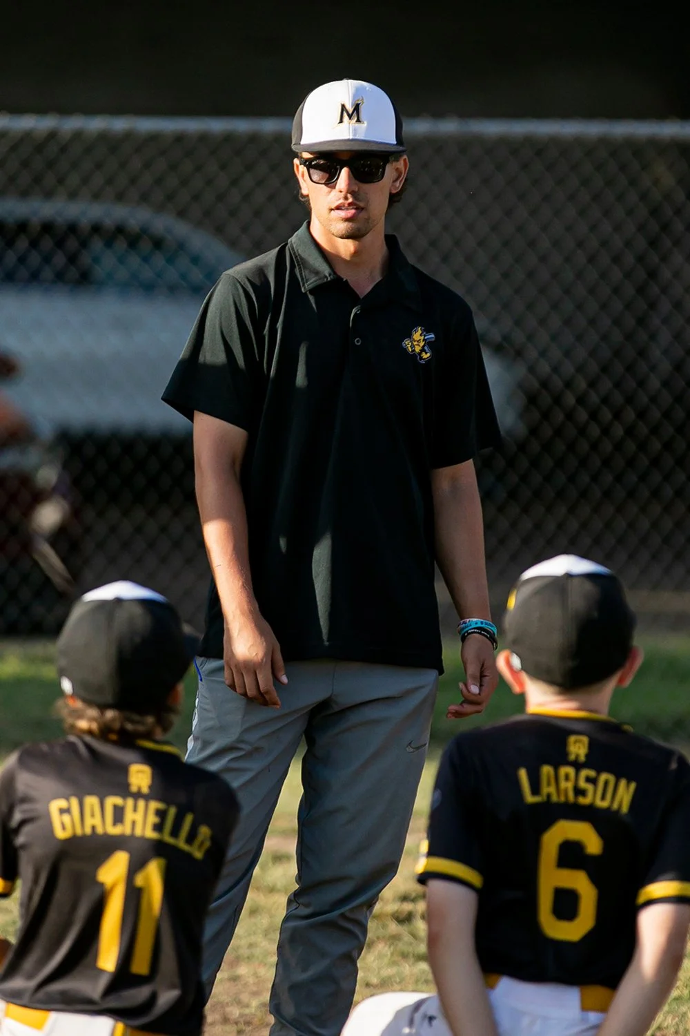 Coach Aaron wearing sunglasses, a black polo shirt with a sports team logo, gray pants, and a baseball cap with a yellow and black design walking on a baseball field.