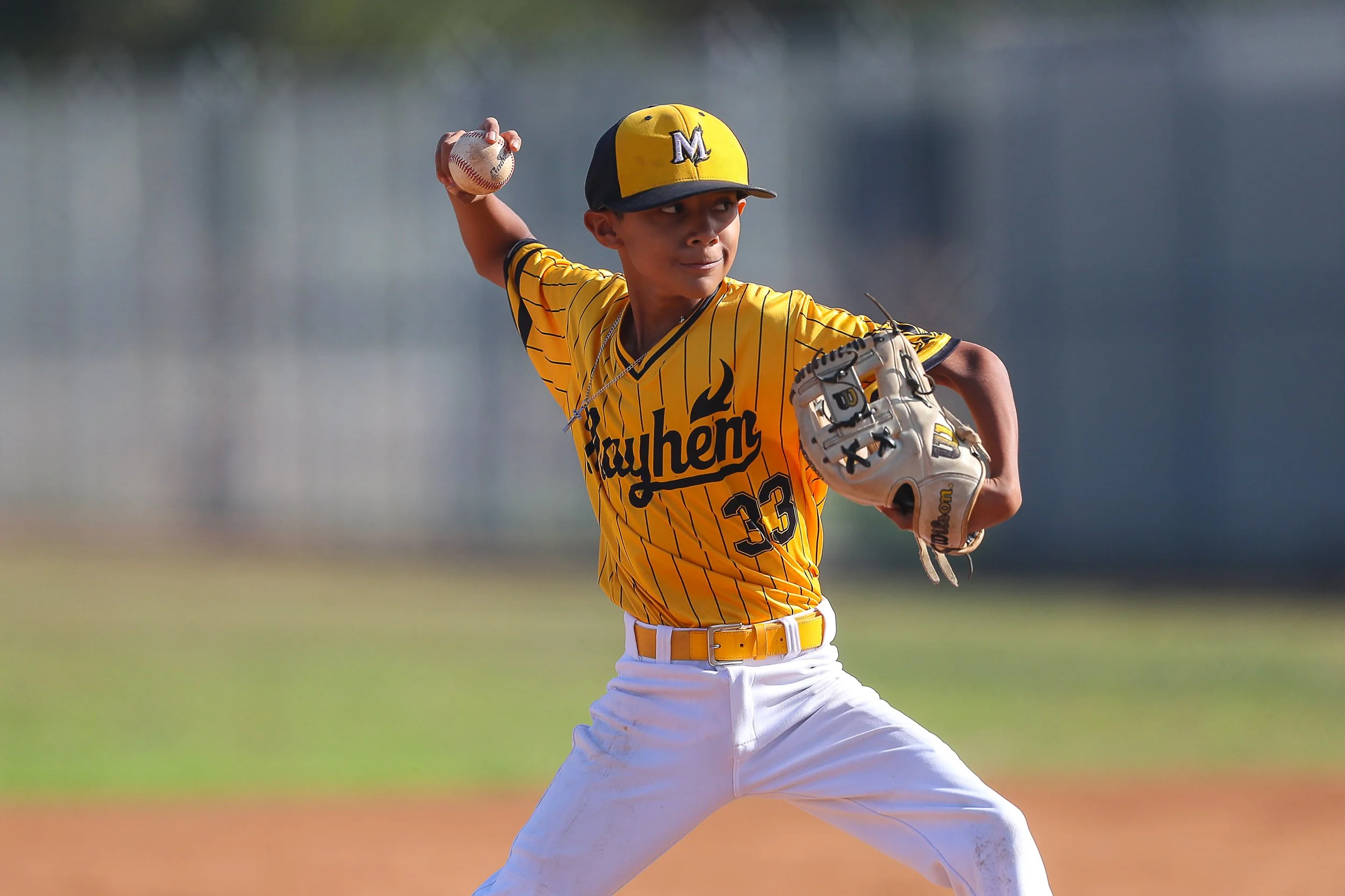 A young baseball player in a yellow and black uniform with the word 'Fauhem' and number 33 on it, wearing a cap, is pitching a baseball on the field.