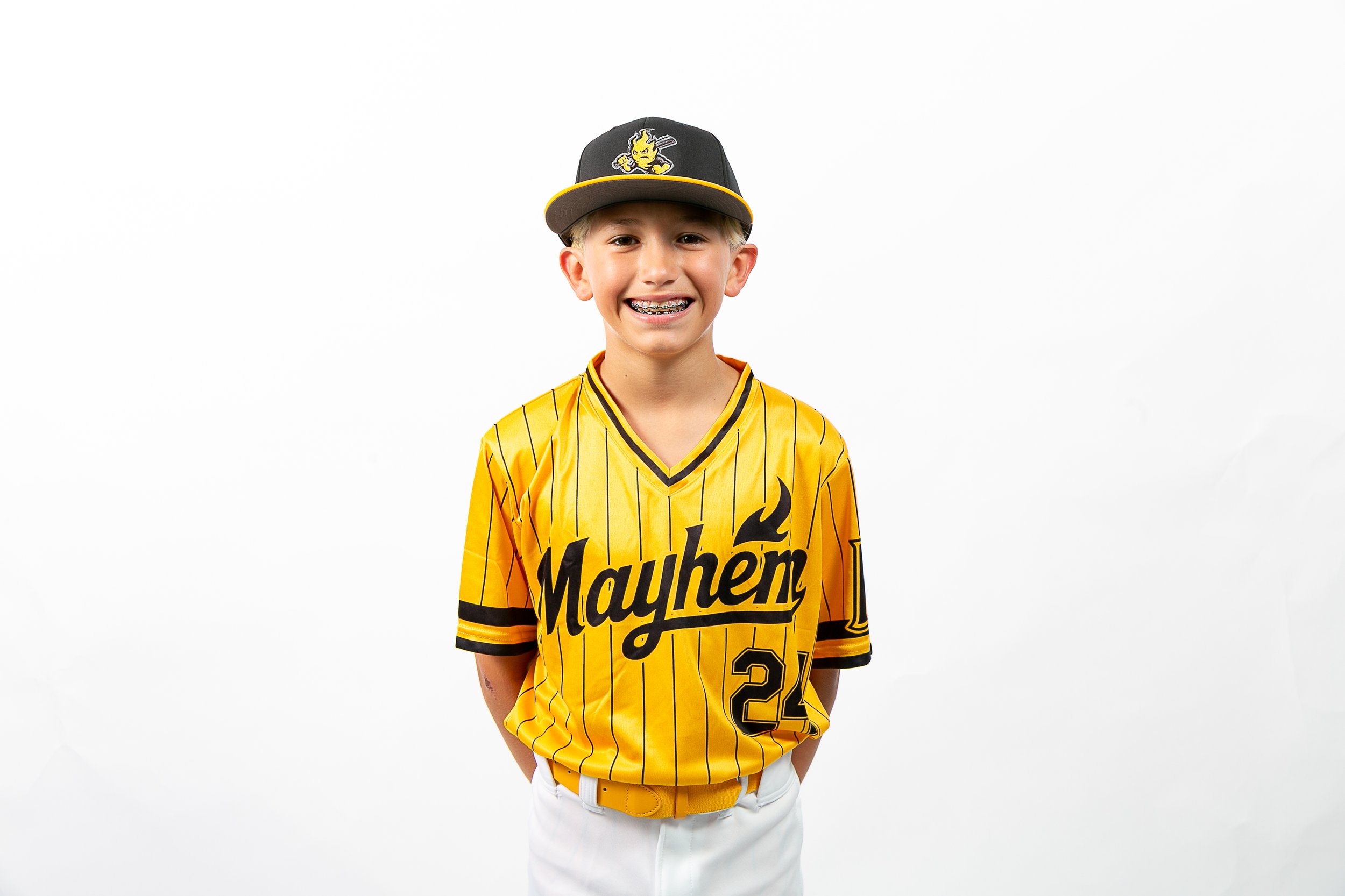 A young boy wearing a yellow and black baseball jersey with the name 'Mayhem' and the number 22, along with a matching black cap with a logo, standing with a smile against a plain white background.