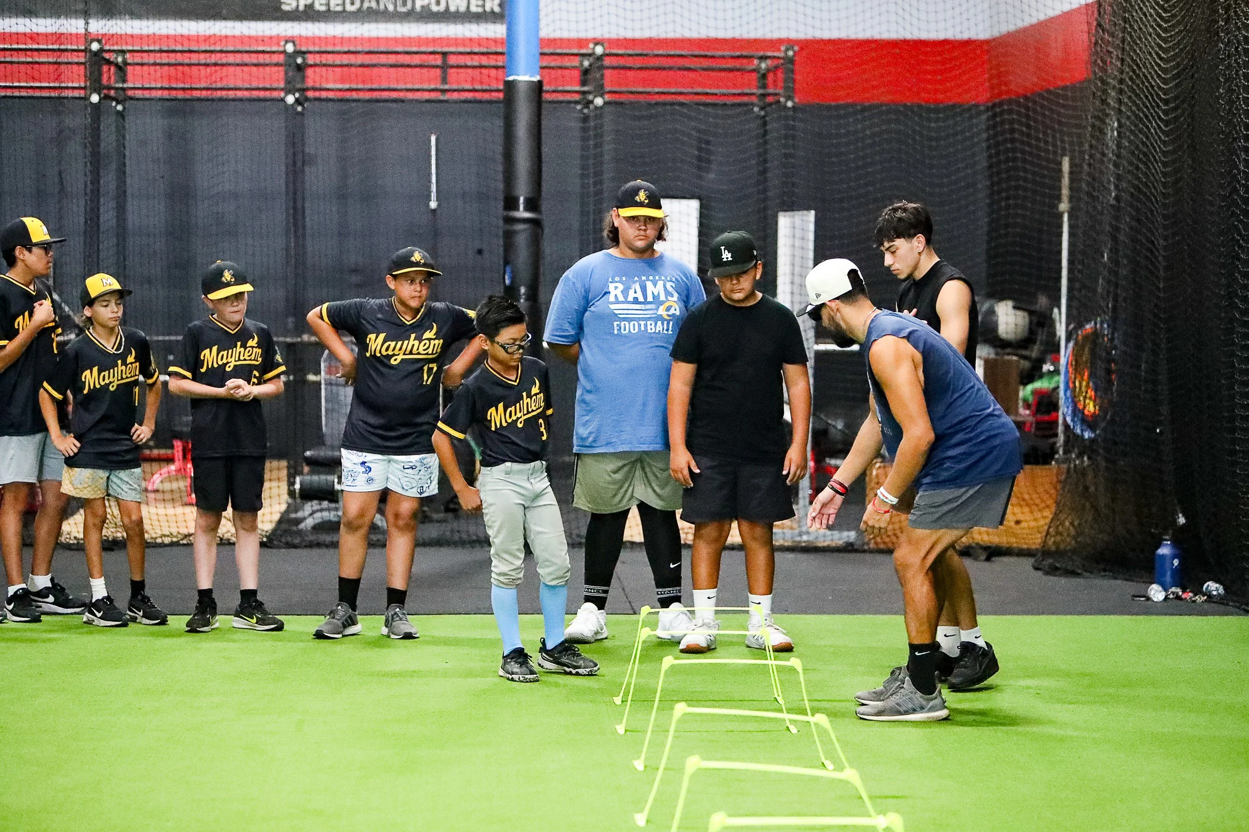 Group of young baseball players and coaches practicing agility drills with hurdles on an indoor field.