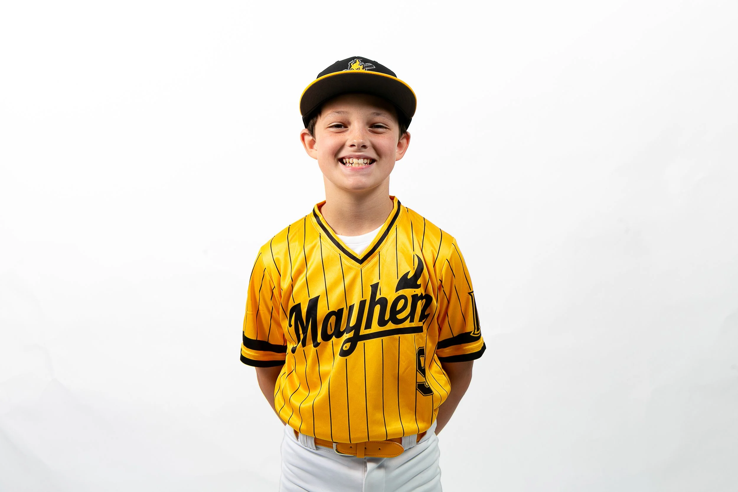 Young boy smiling, wearing a yellow sports jersey with 'Mayhem' written on it, a black cap with yellow accents, and white pants against a plain white background.